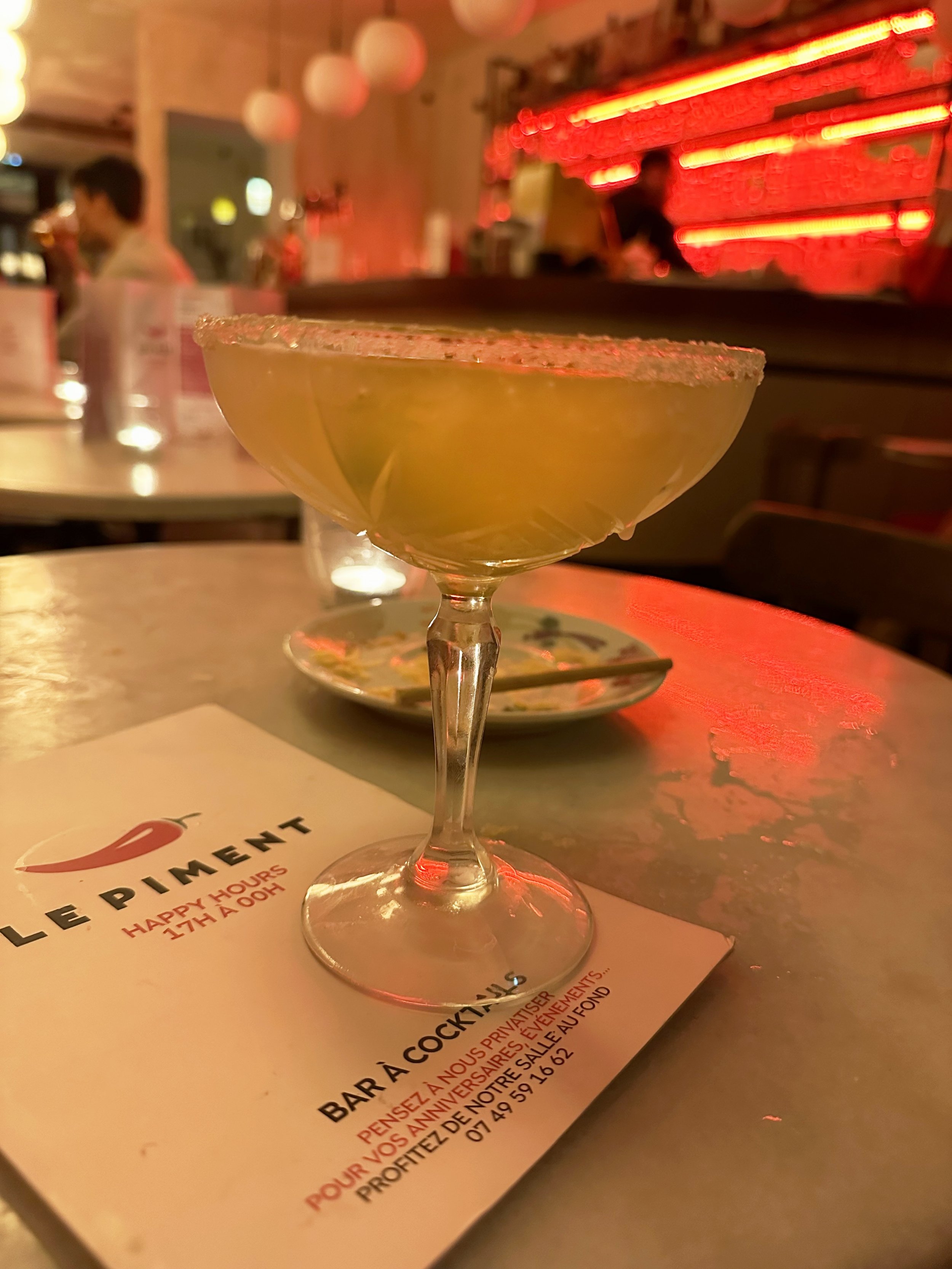 A cocktail glass with a salted rim and a yellow drink on a table in a bar or restaurant with red neon lights, a menu, and a person in the background.