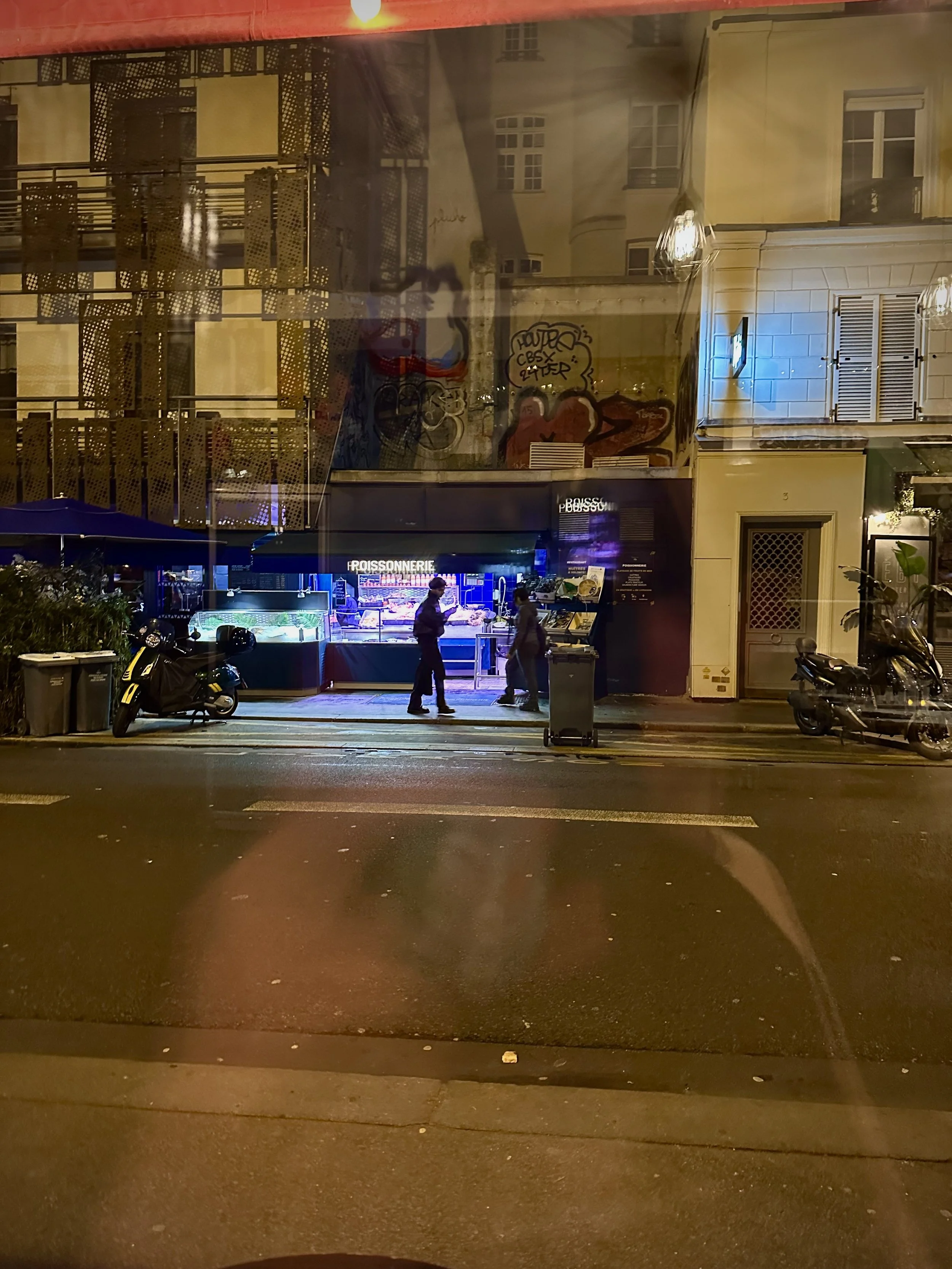 Nighttime street scene with two people standing outside a brightly lit bakery or market stall, with graffiti on the wall behind them, two parked scooters on each side, trash bins, and a reflection of building windows on the glass.