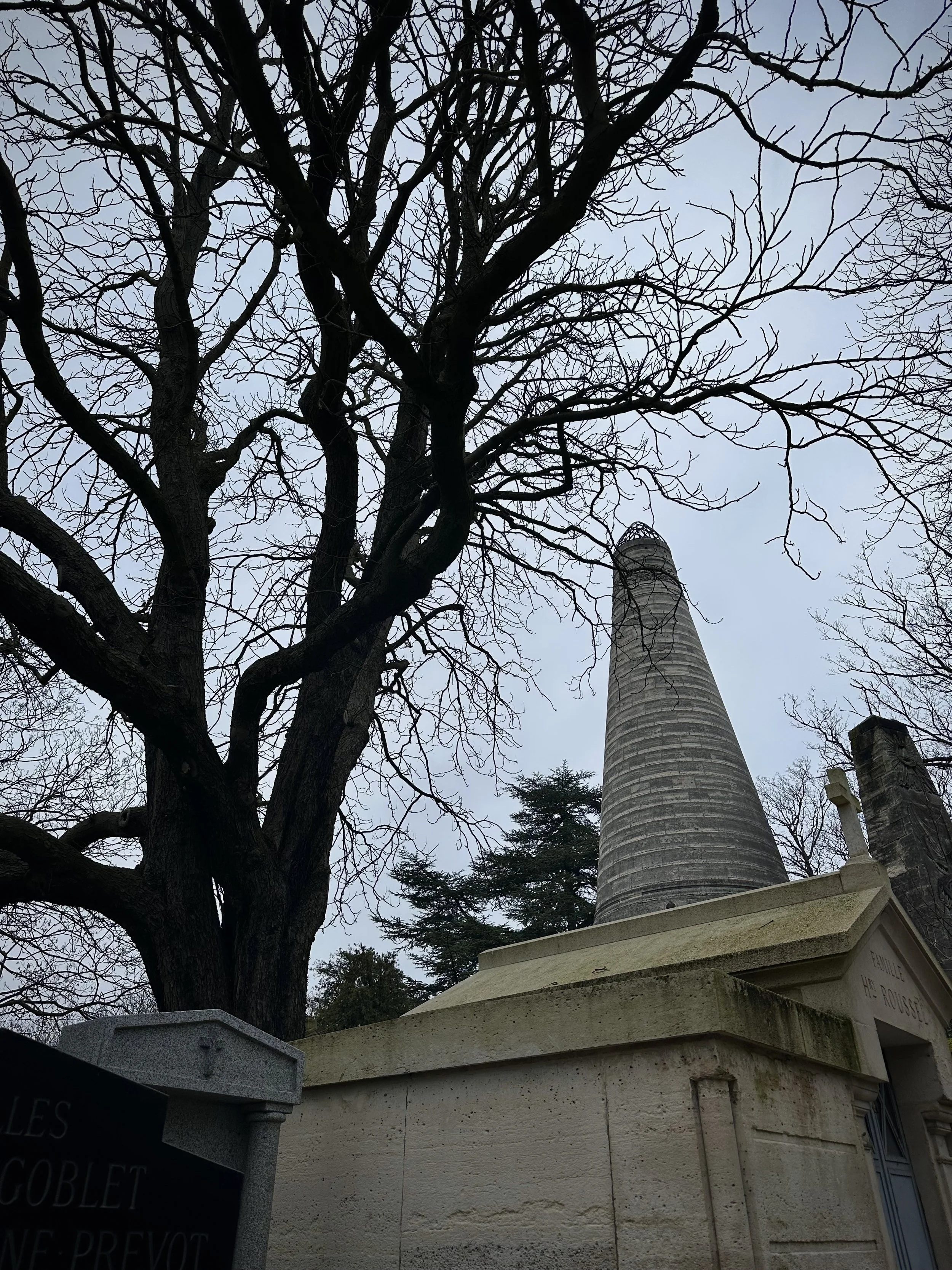 A tall, conical stone chimney next to a leafless tree with numerous branches, and a stone mausoleum with a plaque, set on a cloudy sky background.