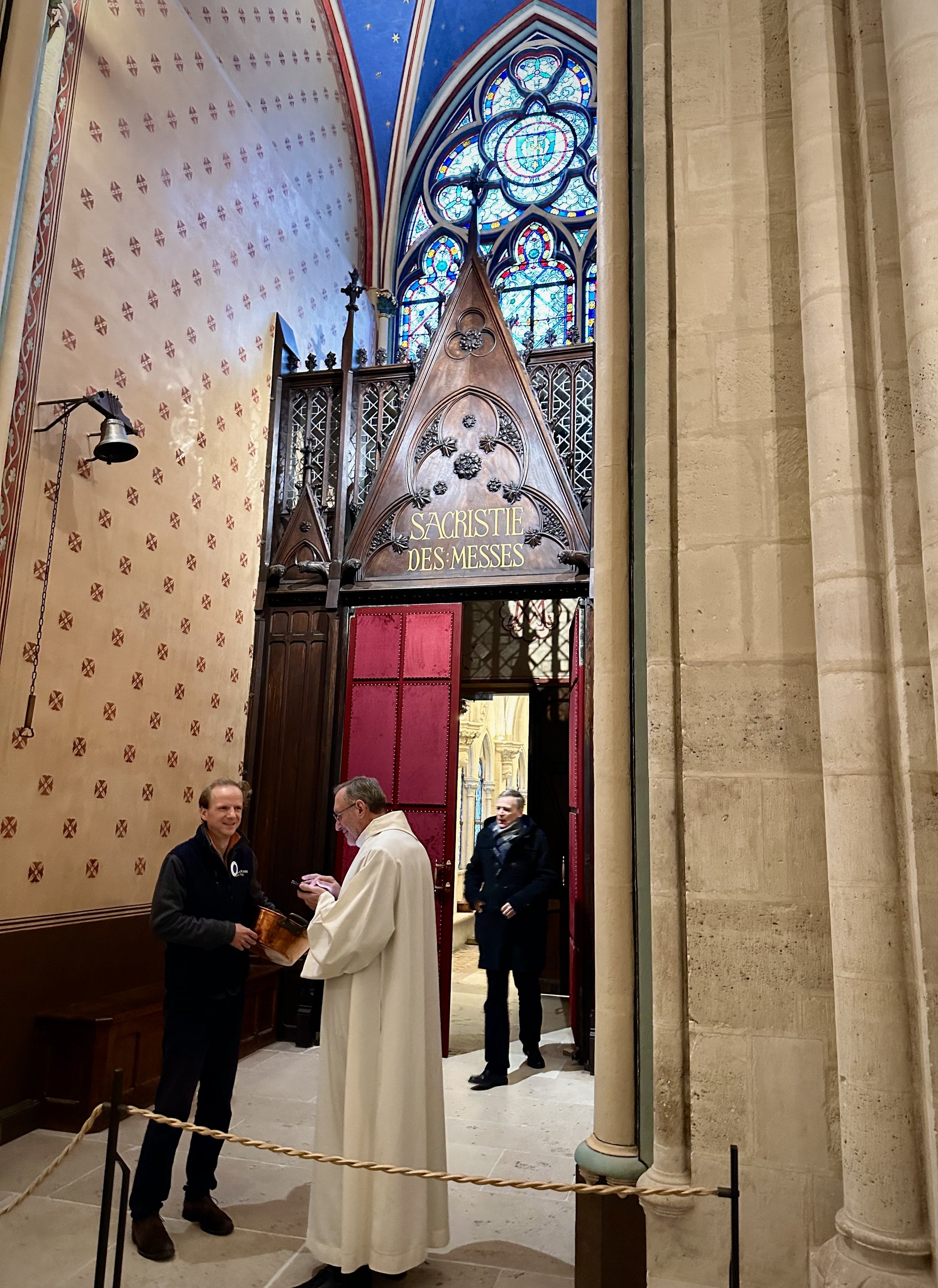 Inside a cathedral with a pointed wooden structure labeled "Sacristie Des Messe," three men are present, with one dressed in religious robes, two others in casual attire, engaging in conversation near a roped-off area. The background features stained glass windows and ornate architectural details.