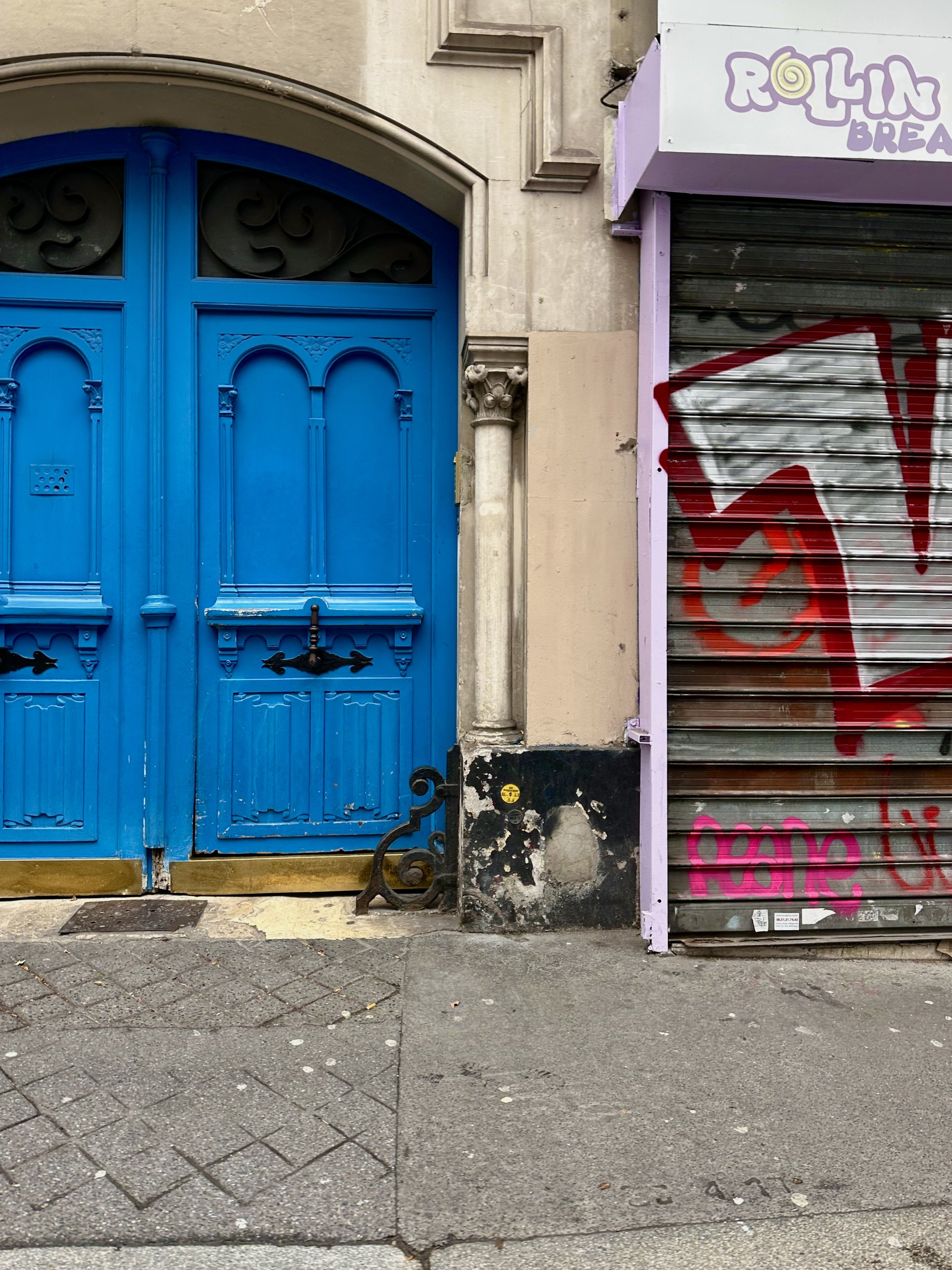 Blue ornate double door with decorative carvings and a black metal handle, adjacent to a wall with graffiti-covered gray metal shutter and a pink and purple sign that reads 'Rollin Bread'.