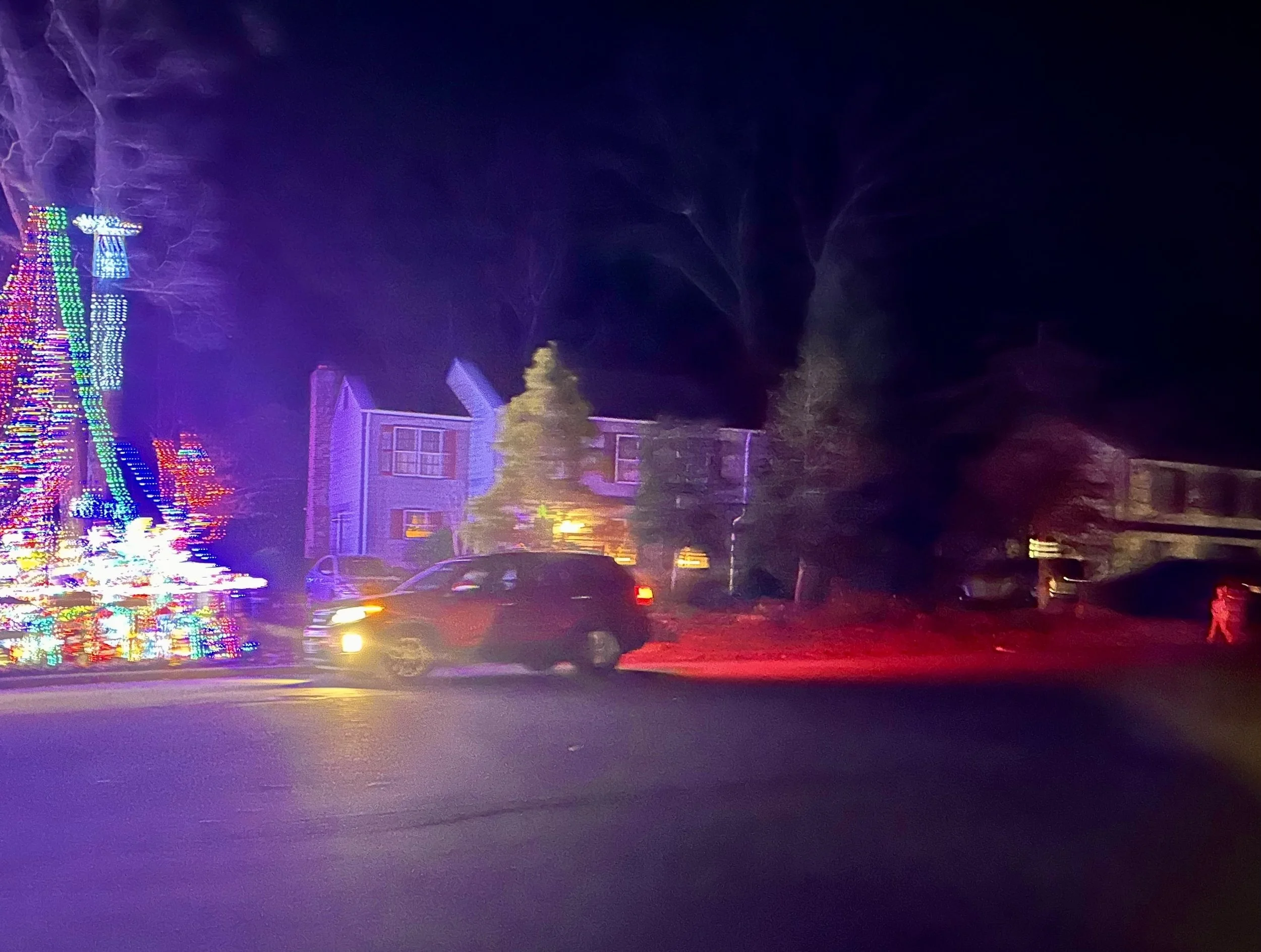 Night scene with colorful Christmas lights on a tree, houses, and nearby area, with a parked car in front and a dark sky in the background.