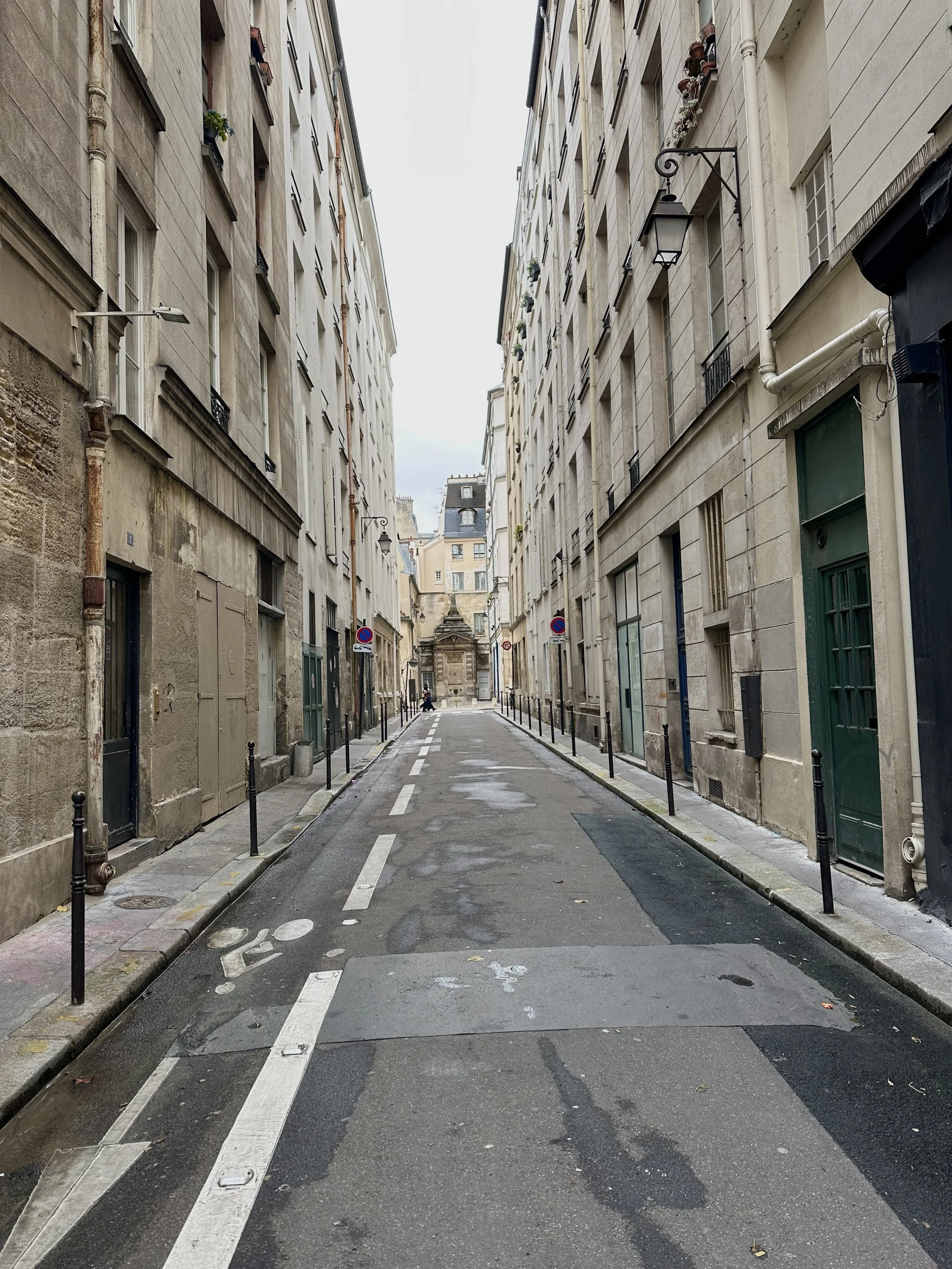 Empty narrow city street with tall buildings on both sides, street markings, and bollards lining the sidewalks, with a building visible in the background.