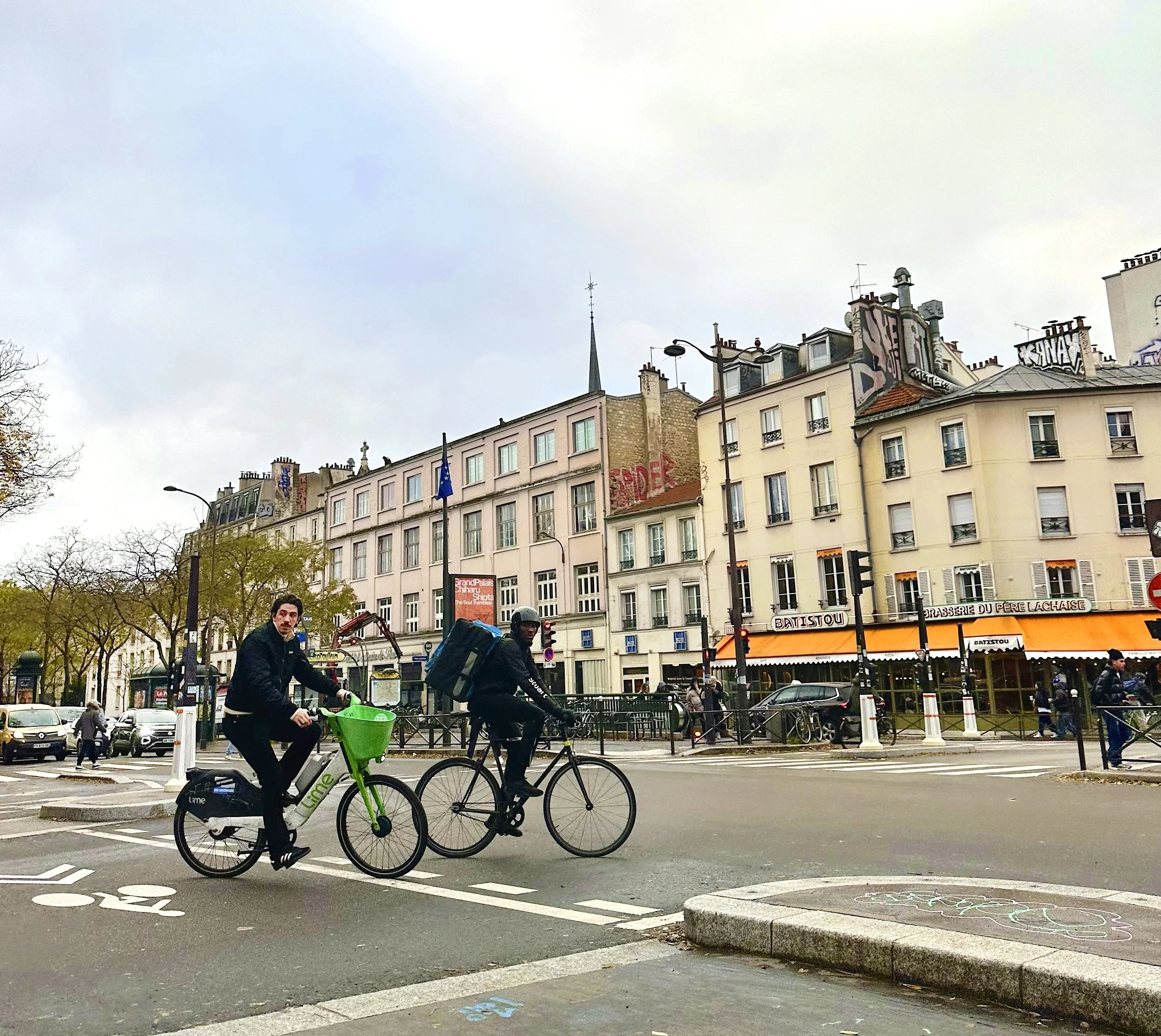 Two men riding bikes at a city intersection with buildings, trees, pedestrians, and cars in the background.