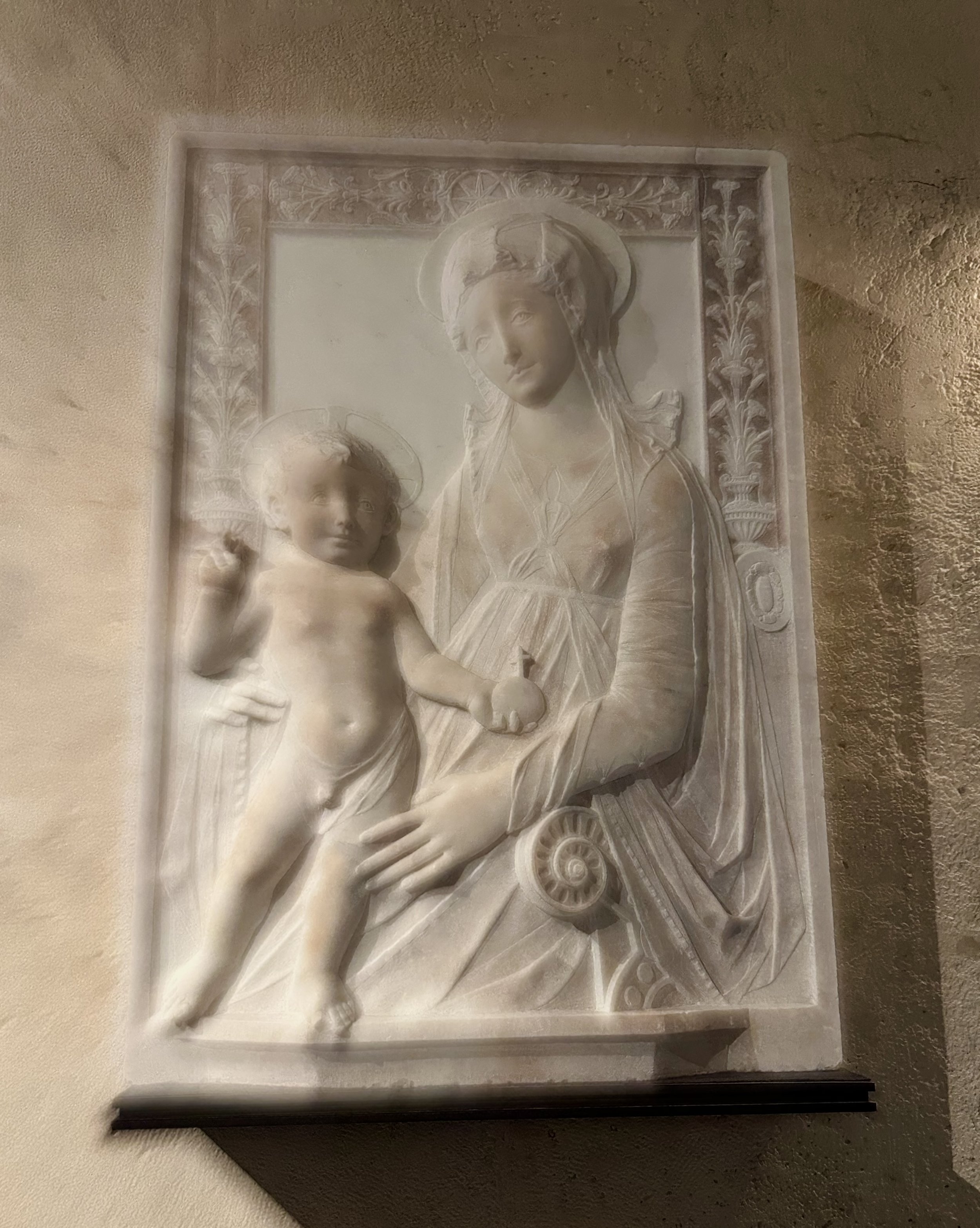 A marble relief sculpture of the Madonna and Child, with the Madonna seated and holding baby Jesus, both with halos, set into a textured stone wall.