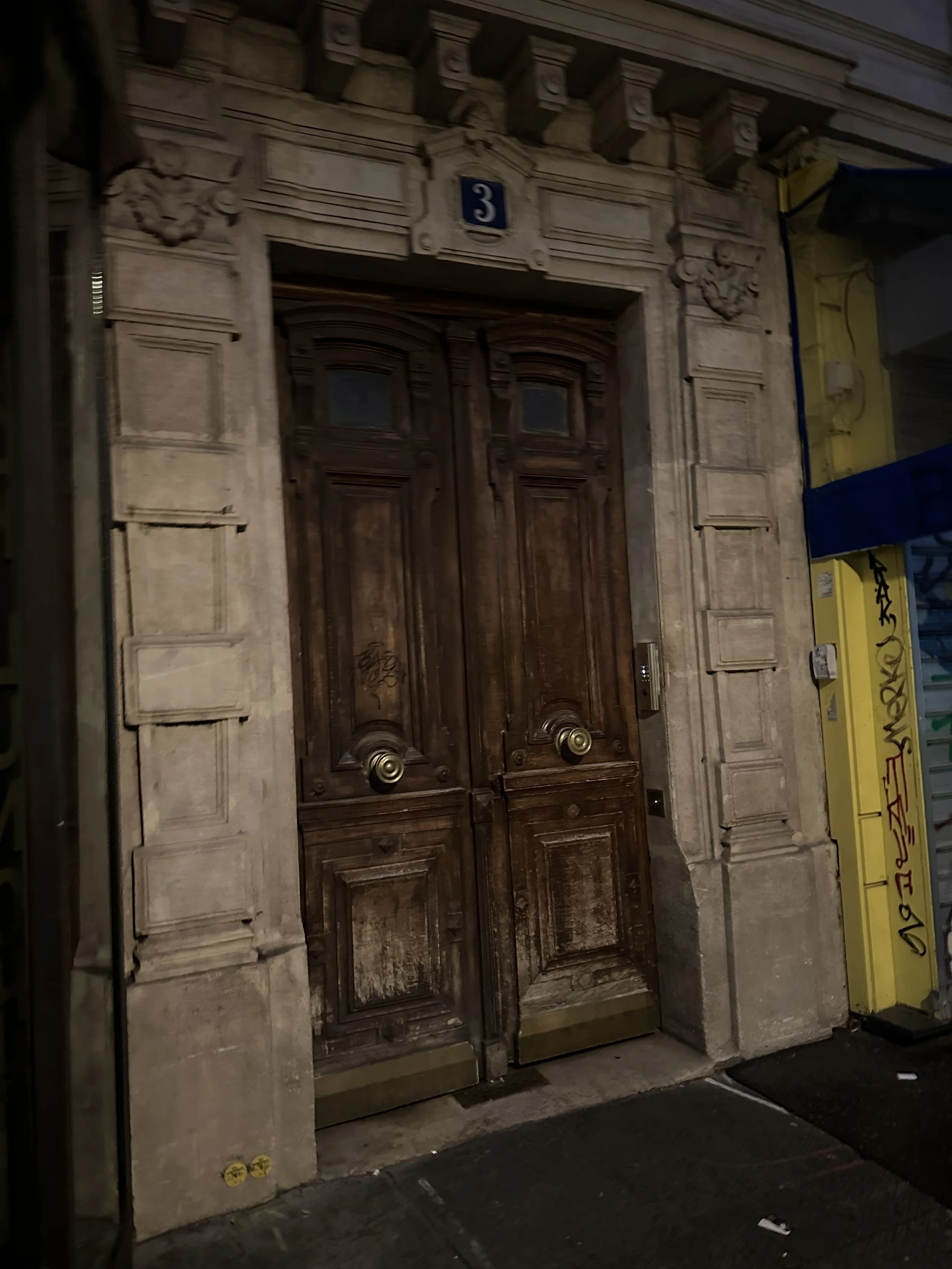 Old double wooden doors with ornate stone frame, number 3 in a small blue plaque above, and graffiti on one door, situated between two narrow yellow and gray walls.
