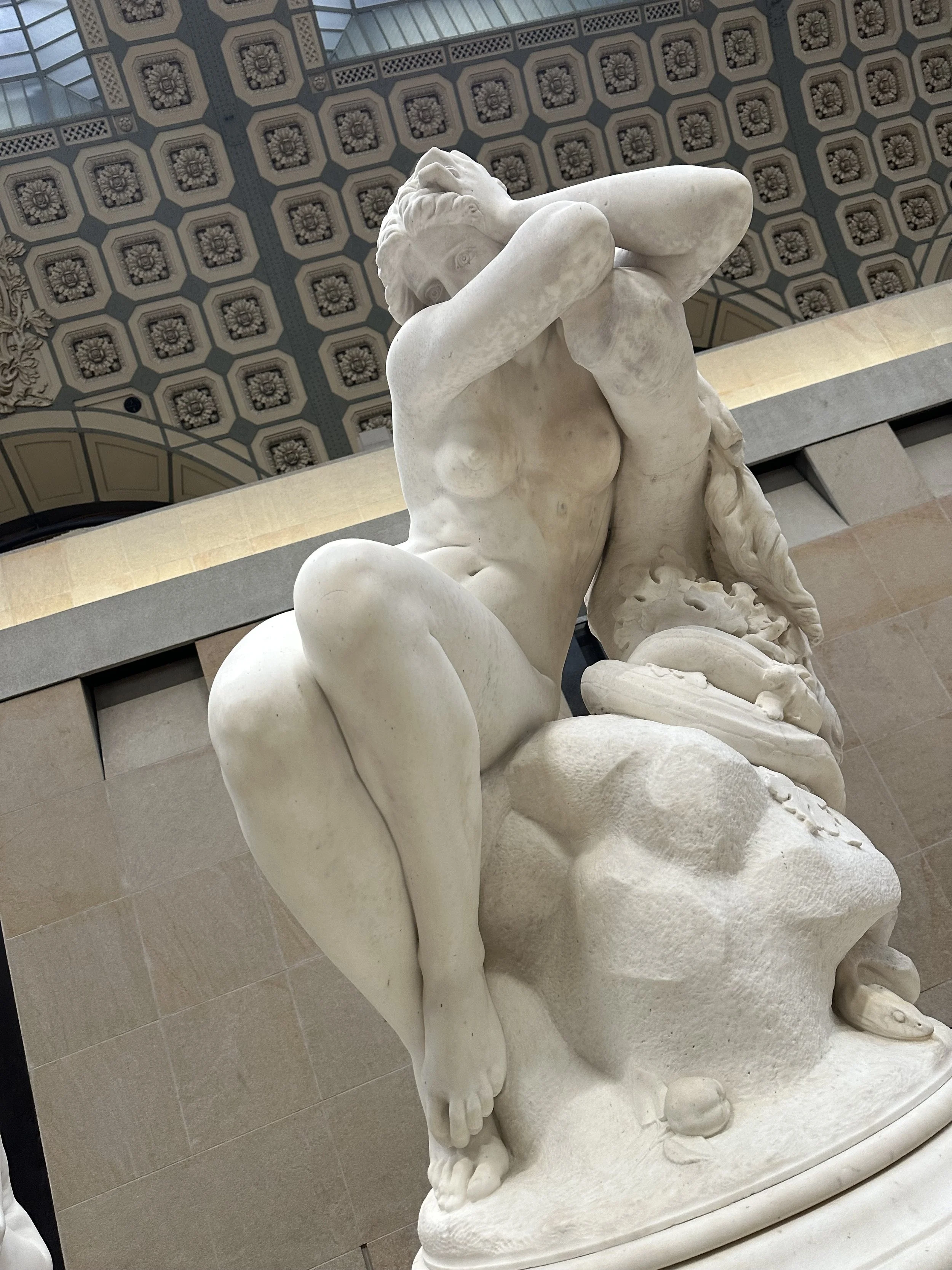 White marble sculpture of a woman covering her face with her arms, seated on a rock, with detailed hair and body features in an ornate indoor setting.