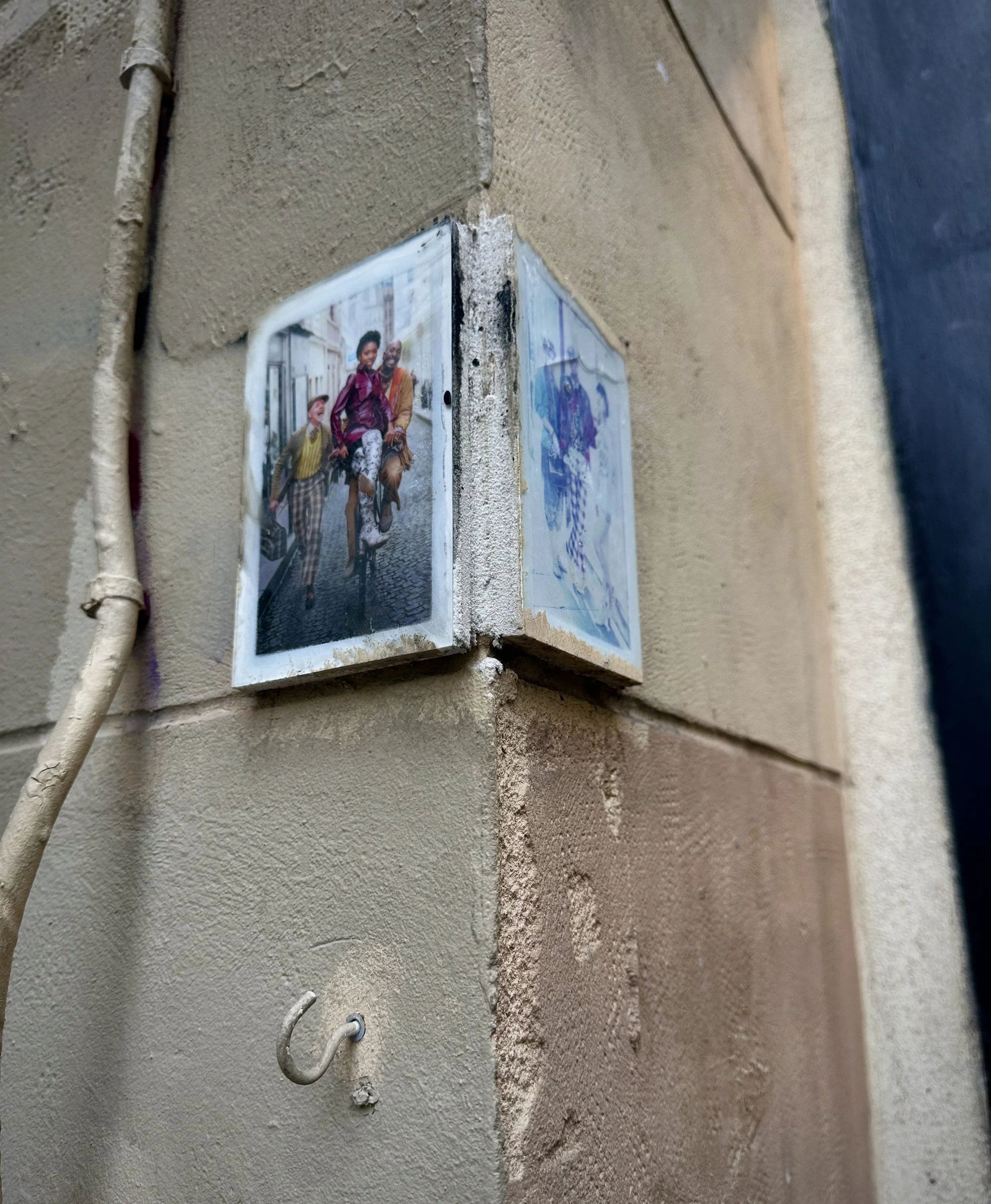Photograph of a corner of a building with two small picture frames attached to the beige-colored brick wall. One frame shows a colorful street scene with people walking.