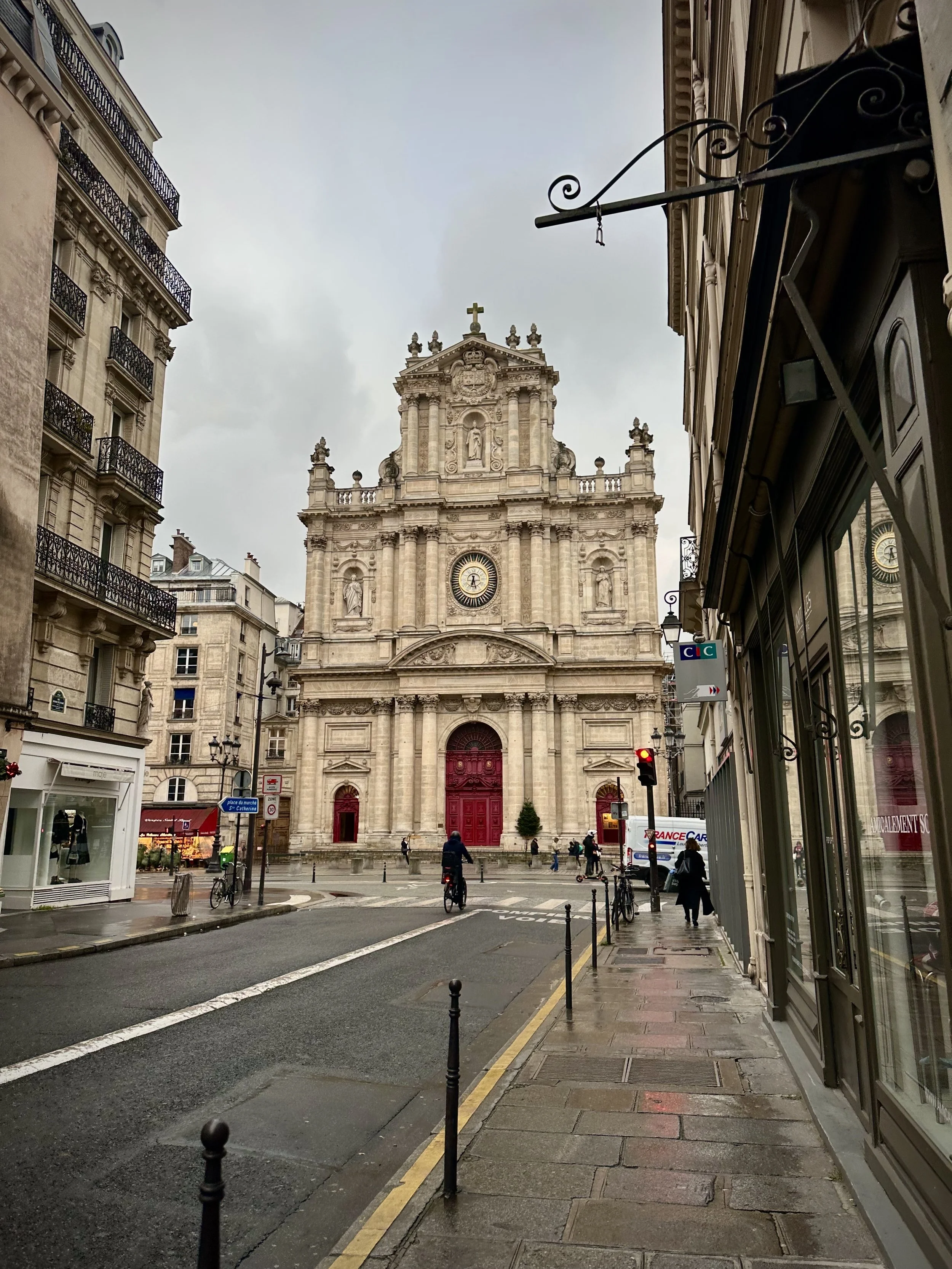 A historic church with ornate architecture, statues, and a cross on top, situated at the end of a city street with people biking and walking on a cloudy day.