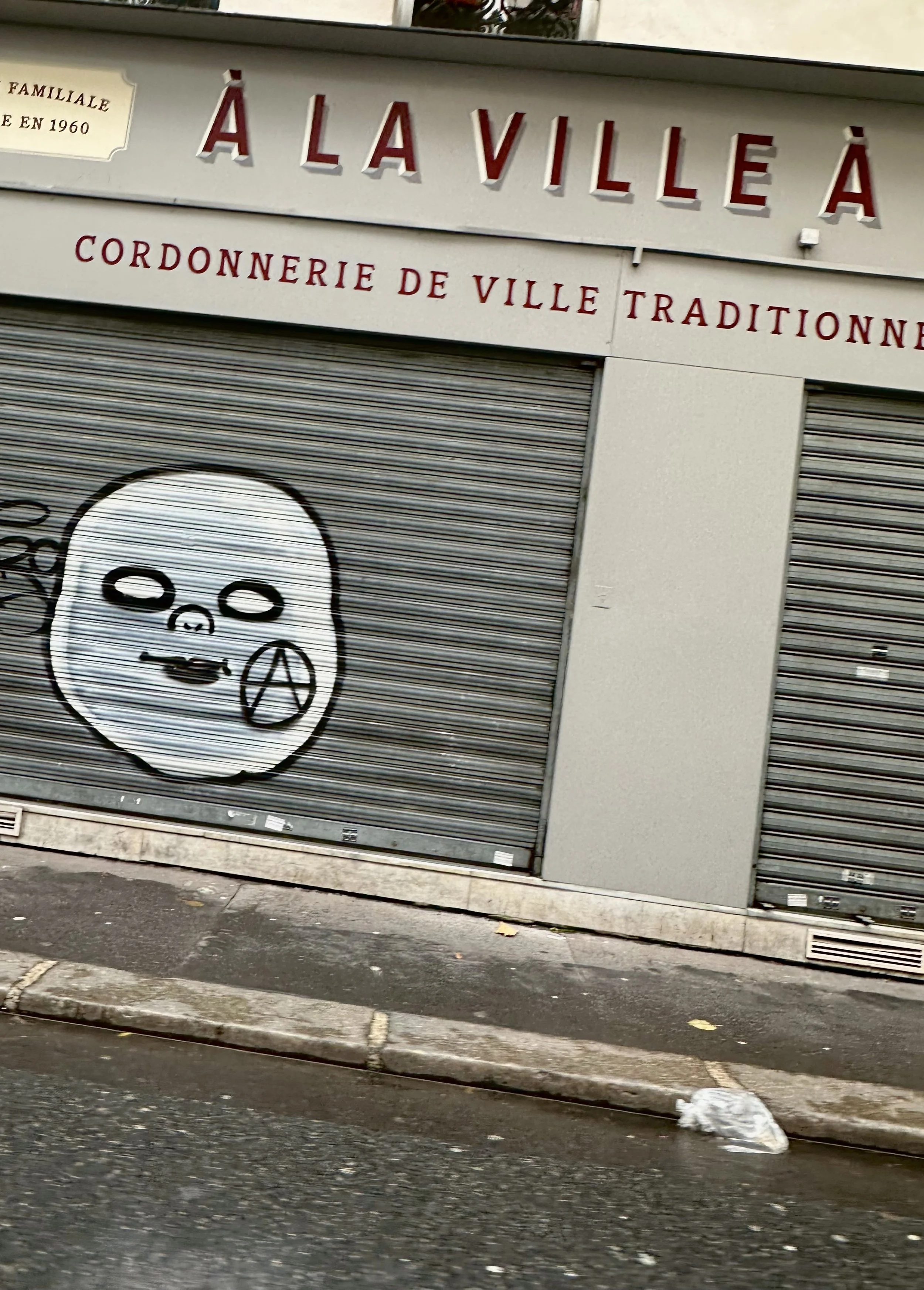 Closed storefront with graffiti of a cartoonish skull face with peace symbol nose on the metal shutter, above French signs for a traditional city butcher shop.