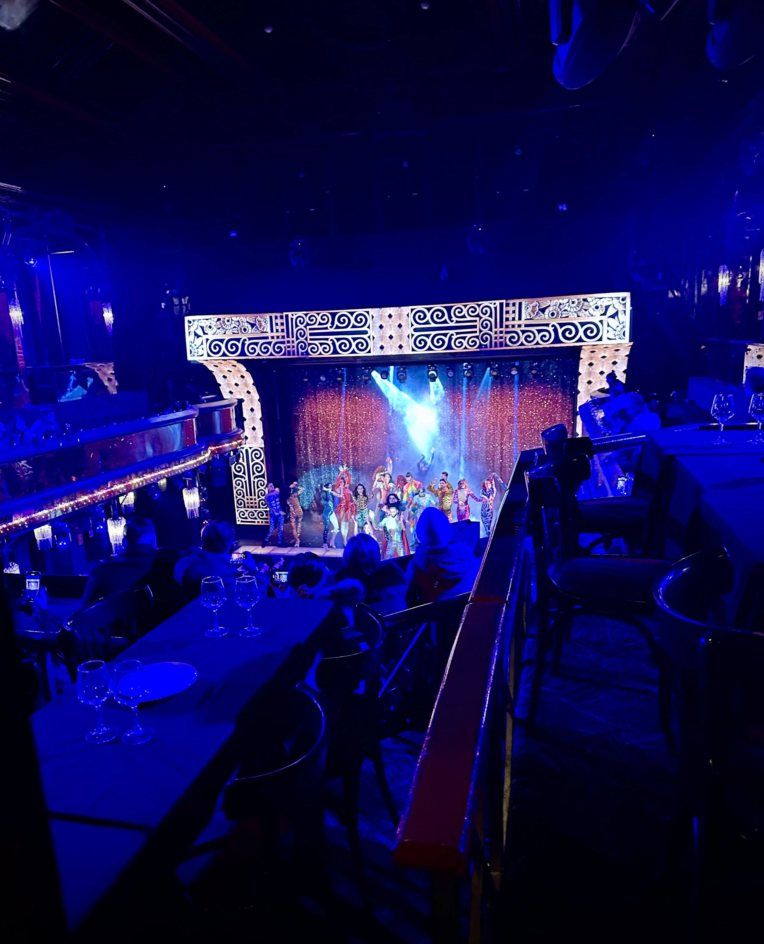 The image shows a vibrant stage performance at a theater with colorful costumes, bright lights, and a decorated backdrop. Audience members are seated at tables watching the show.
