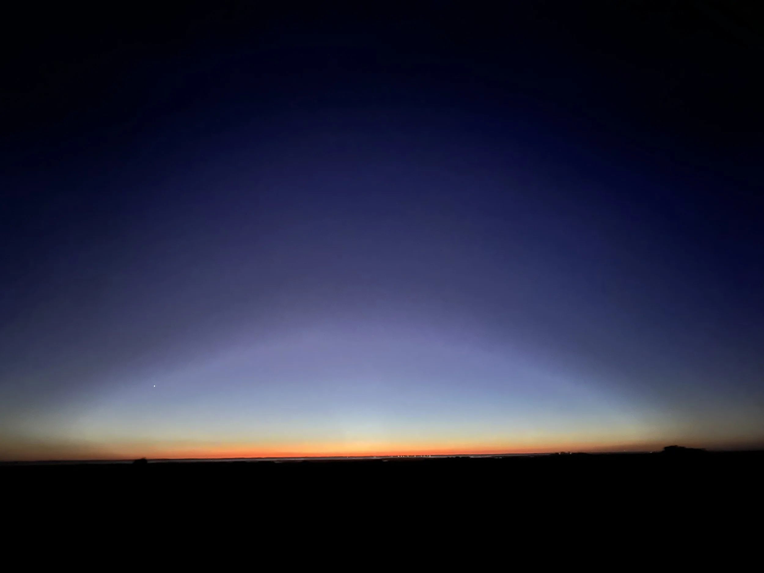 A sunset scene with a dark horizon, a faint star or planet in the sky, and a gradient of colors from orange and yellow near the horizon to deep blue and black overhead.