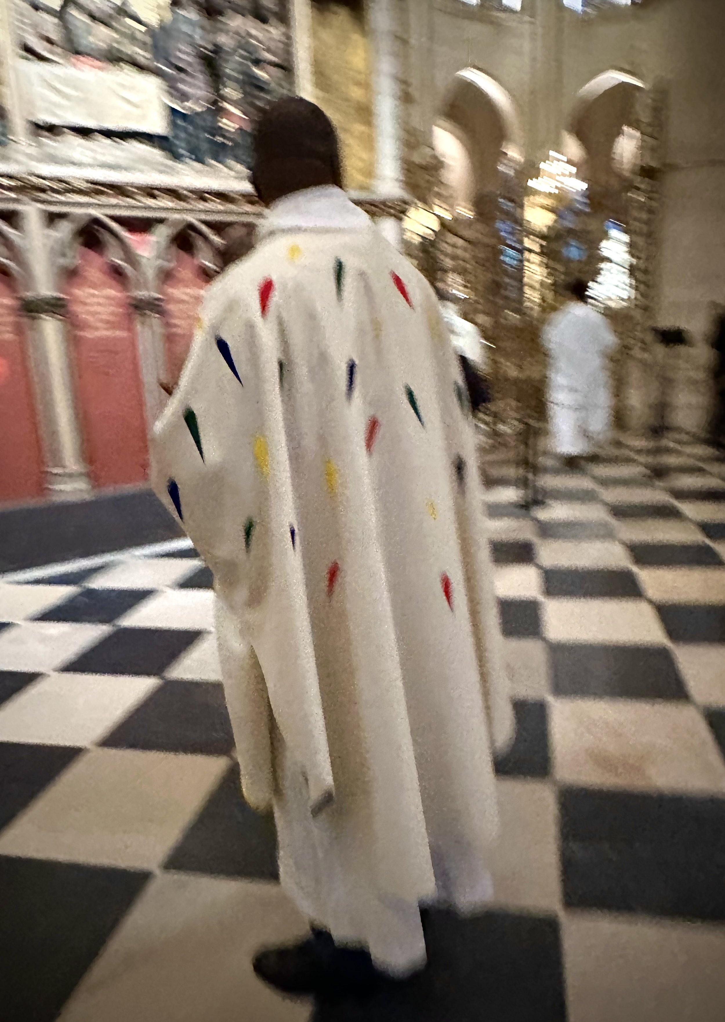 A person wearing a white robe decorated with multicolored candle-shaped patterns inside a church with black and white checkered floor and ornate architecture.