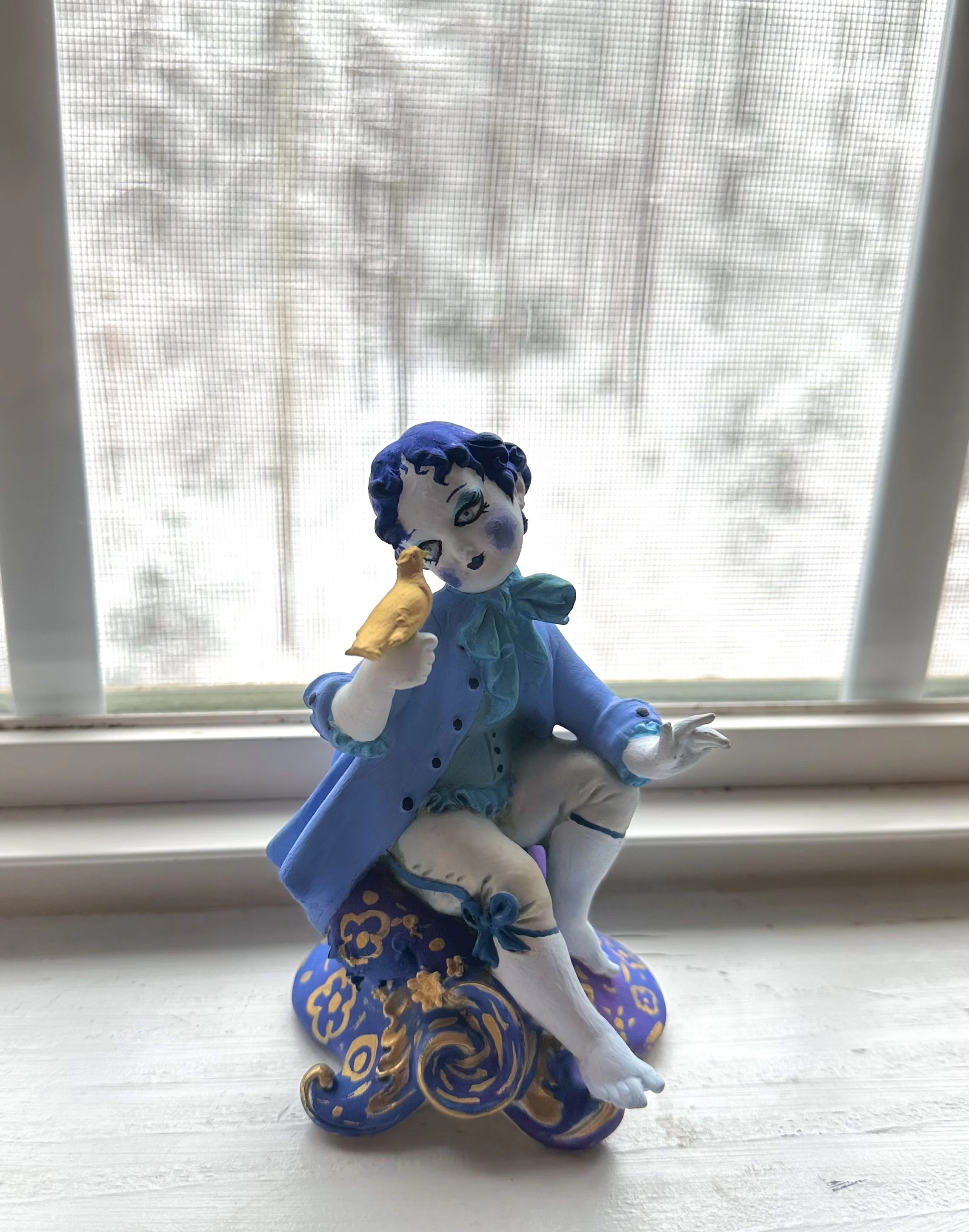Porcelain figurine of a boy with blue hair, wearing a blue coat, sitting on a decorative blue and gold swirl base, holding a yellow bird near his face, placed on a windowsill with a snowy scene outside.