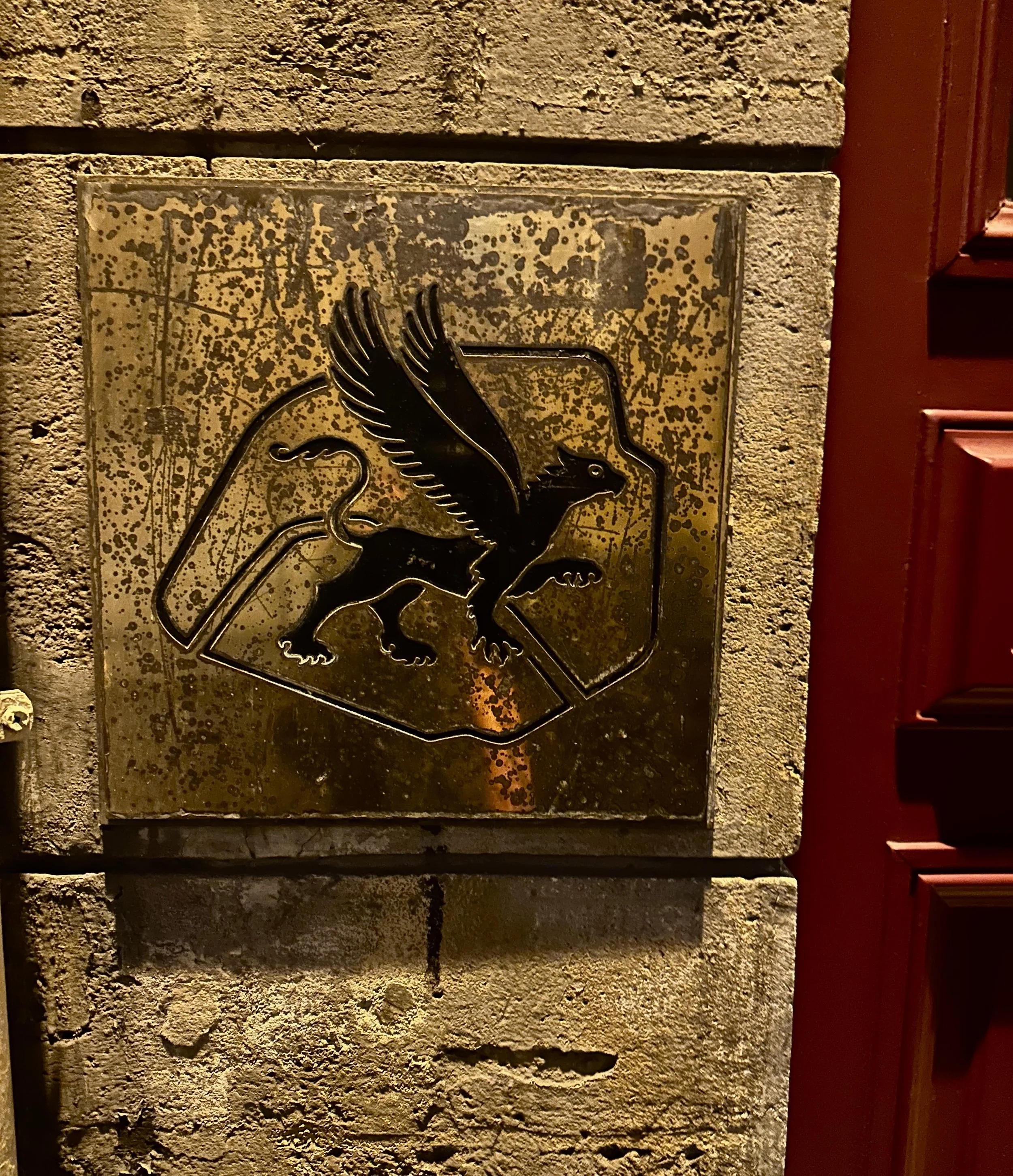 Metal plaque on stone wall featuring a winged lion with a raised paw, outlined in black.