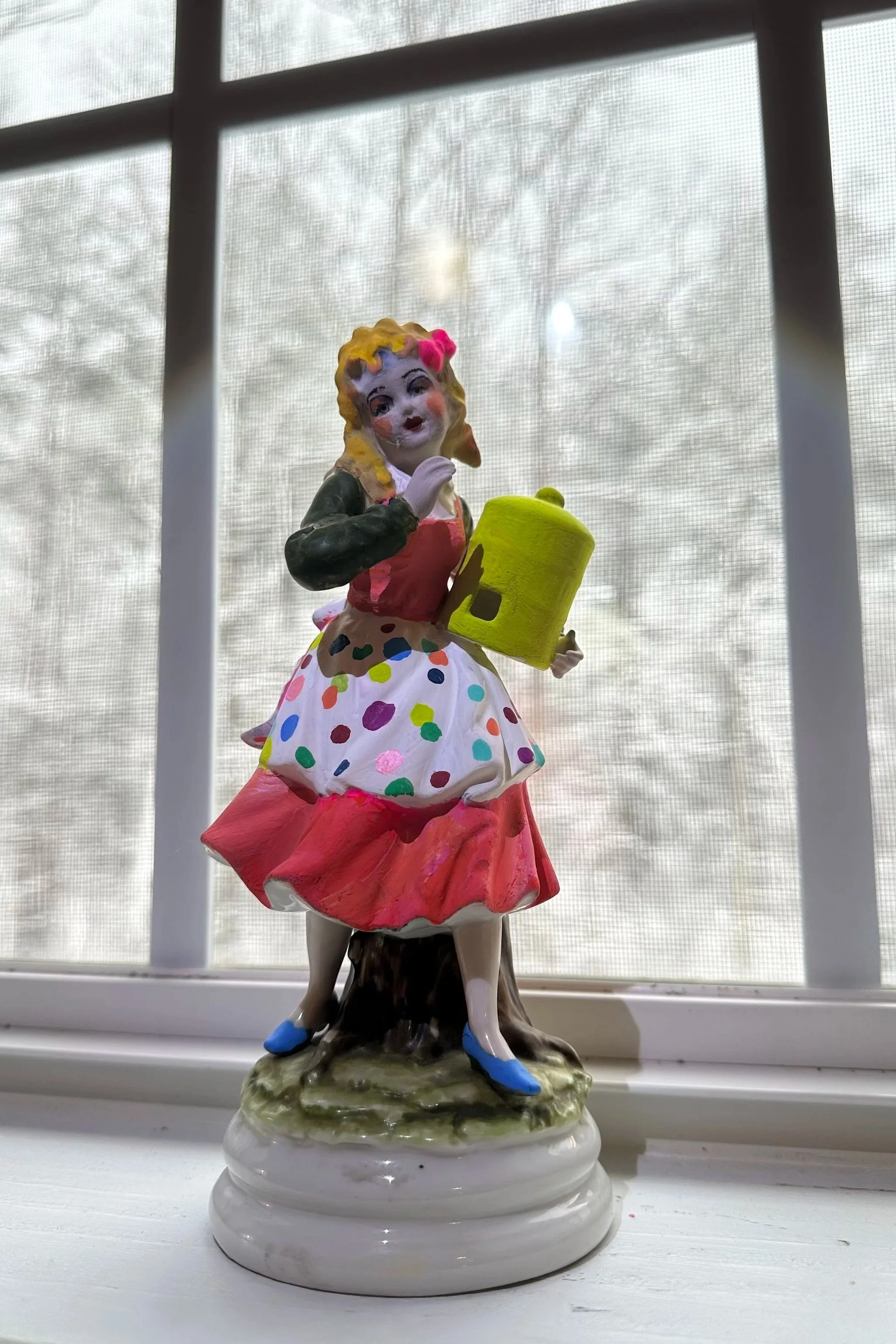 Colorful ceramic figurine of a girl in a polka dot dress holding a yellow book, standing on a decorated base near a window with a snowy landscape visible outside.