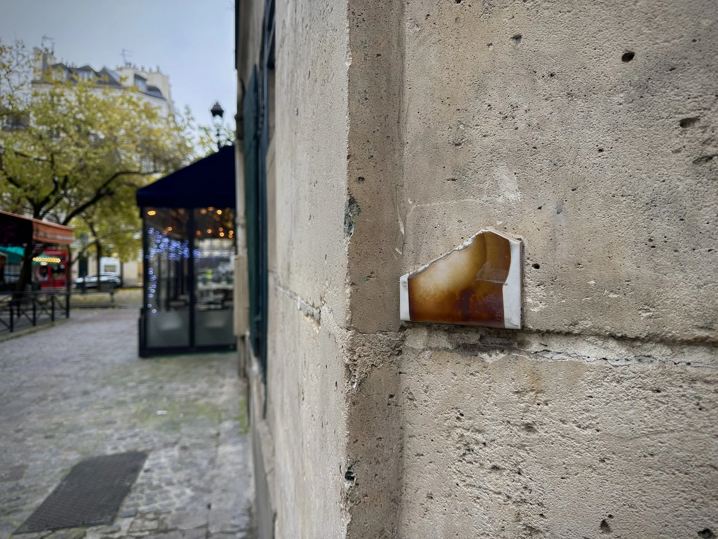 Close-up of a small, tattered ceramic sign with a brown and white glazed surface attached to a rough, beige stone wall in an outdoor urban setting, with blurred trees, buildings, and a cafe or shop with string lights in the background.