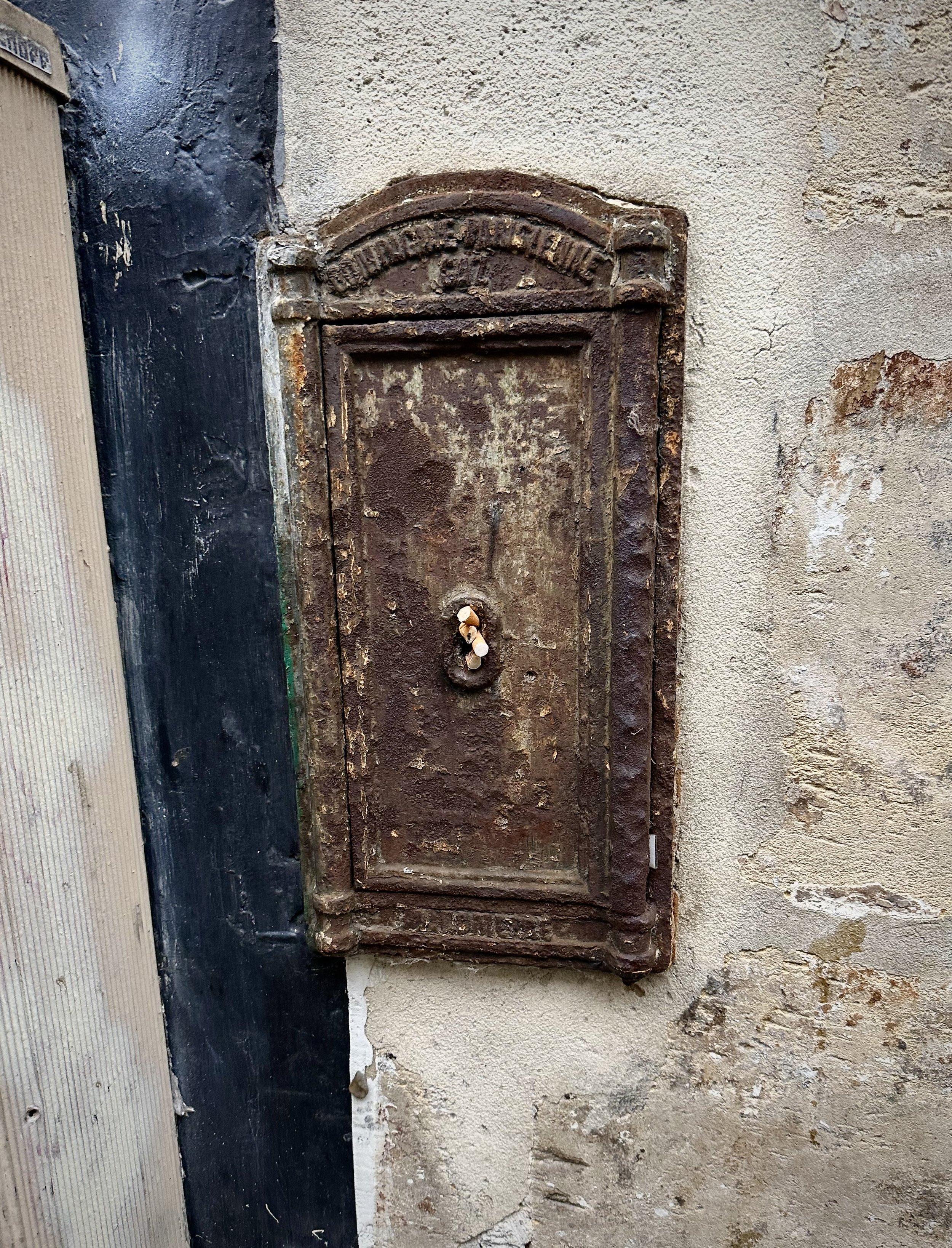 An old, rusted metal mailbox attached to an aged, textured wall with peeling paint, featuring an arched top and a handle with a lock.
