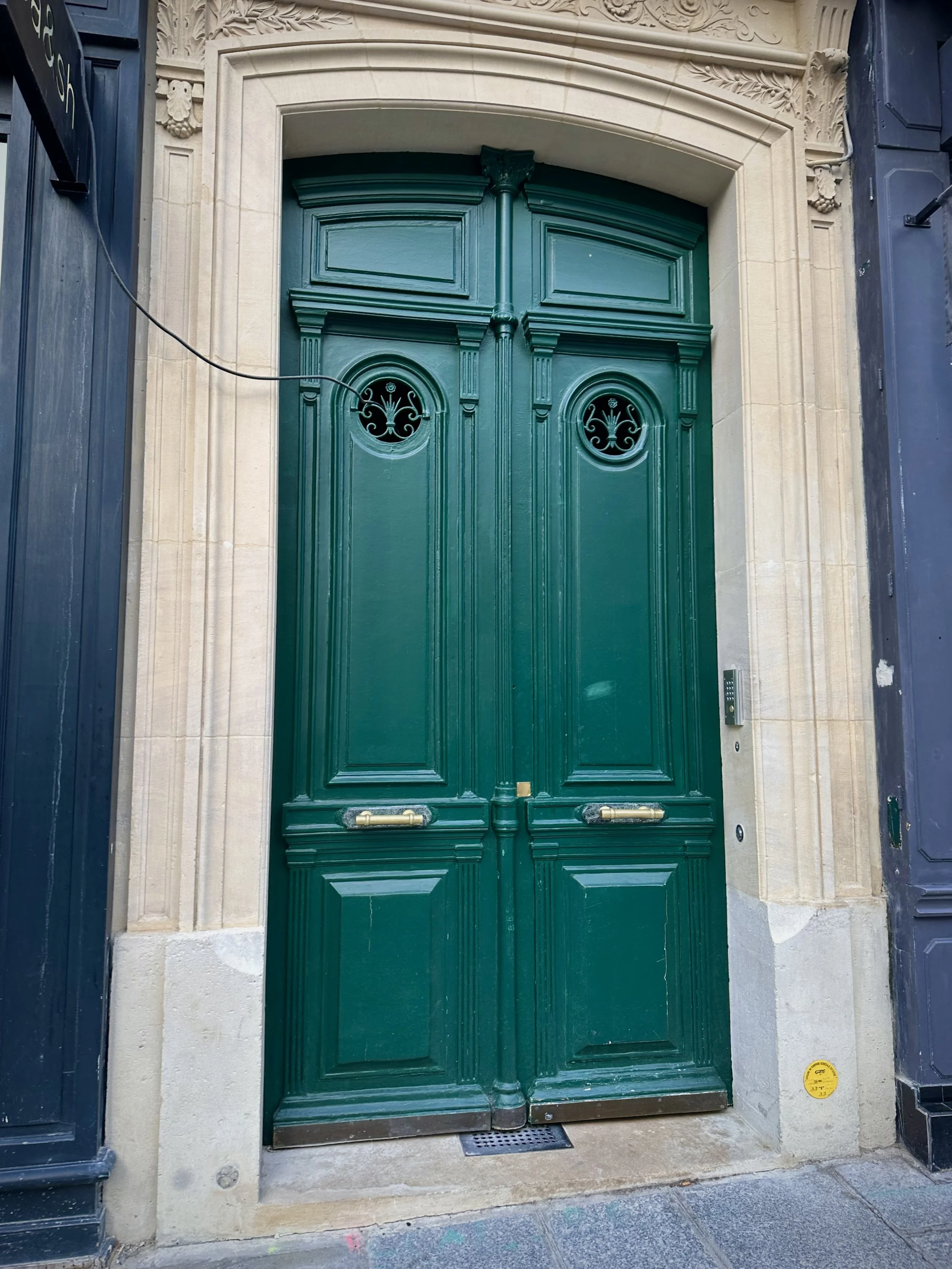 Green double door with decorative round windows, set in stone framing, with a security camera and intercom to the side.