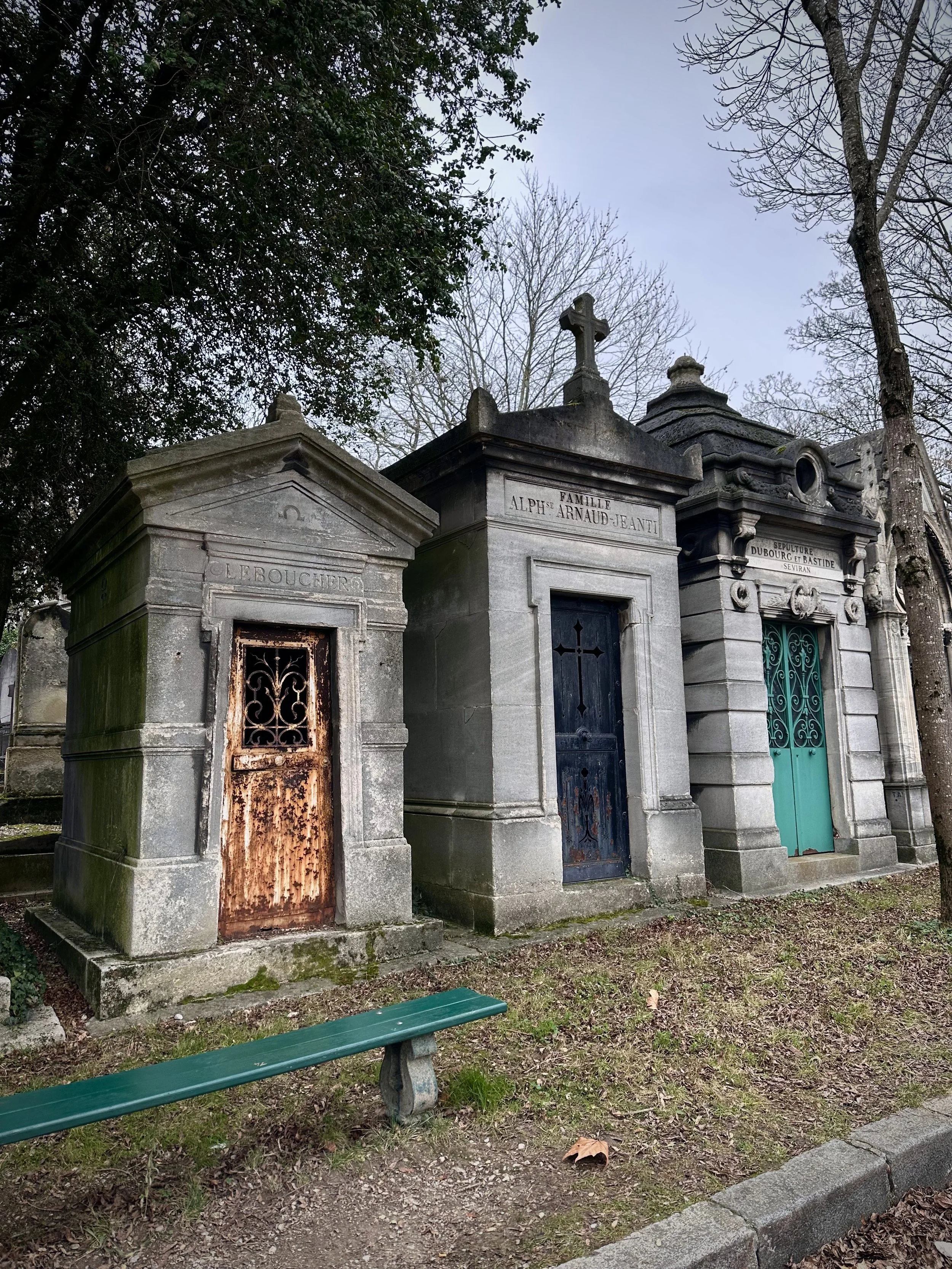 Three old mausoleum tombs in a cemetery, made of stone with metal doors. One is rusted, and the others are painted blue and turquoise. There are trees around and a green bench in front.