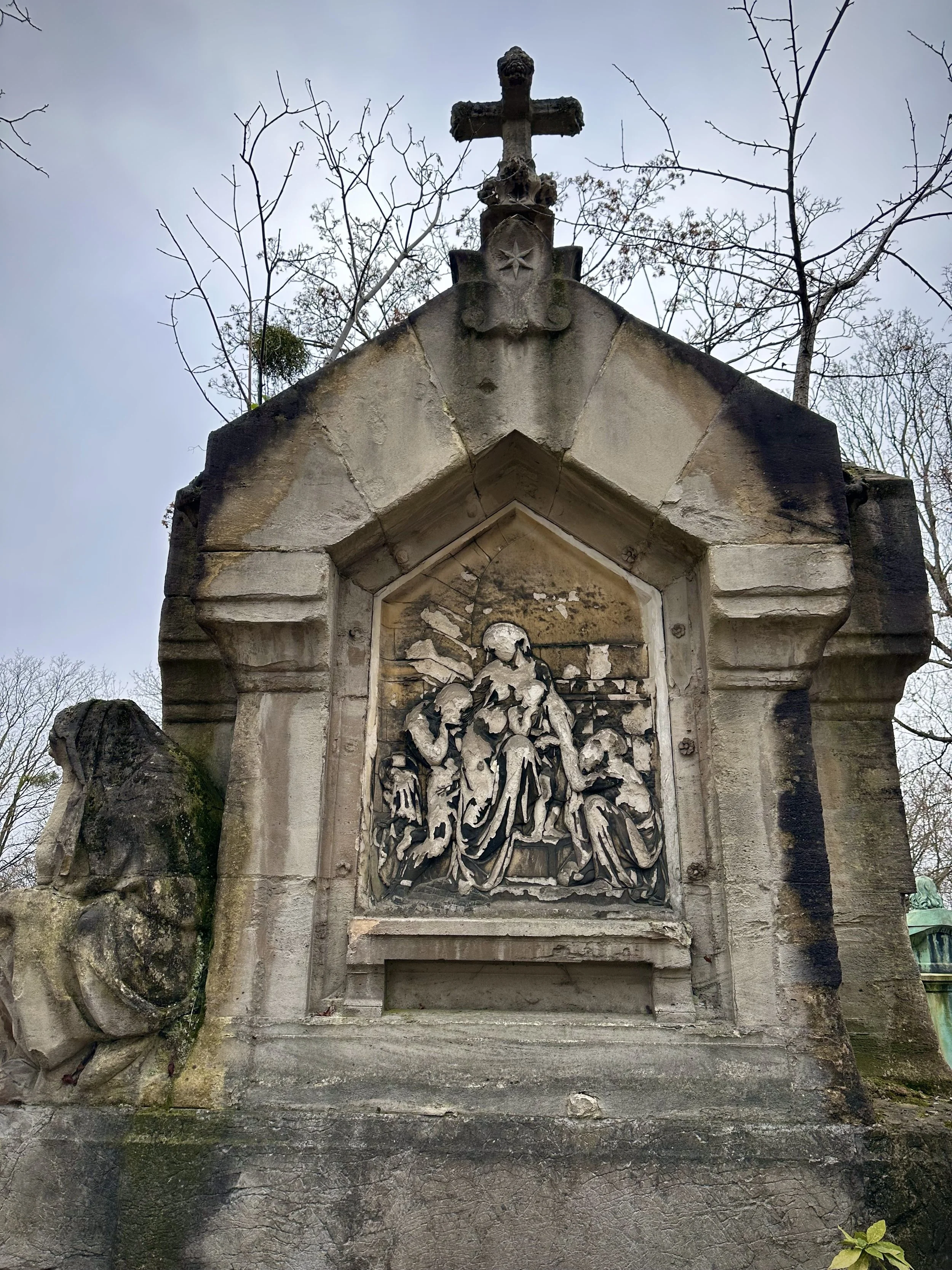 A stone religious monument with a carved relief scene of the Virgin Mary and children, topped with a cross, in an outdoor setting with bare trees in the background.