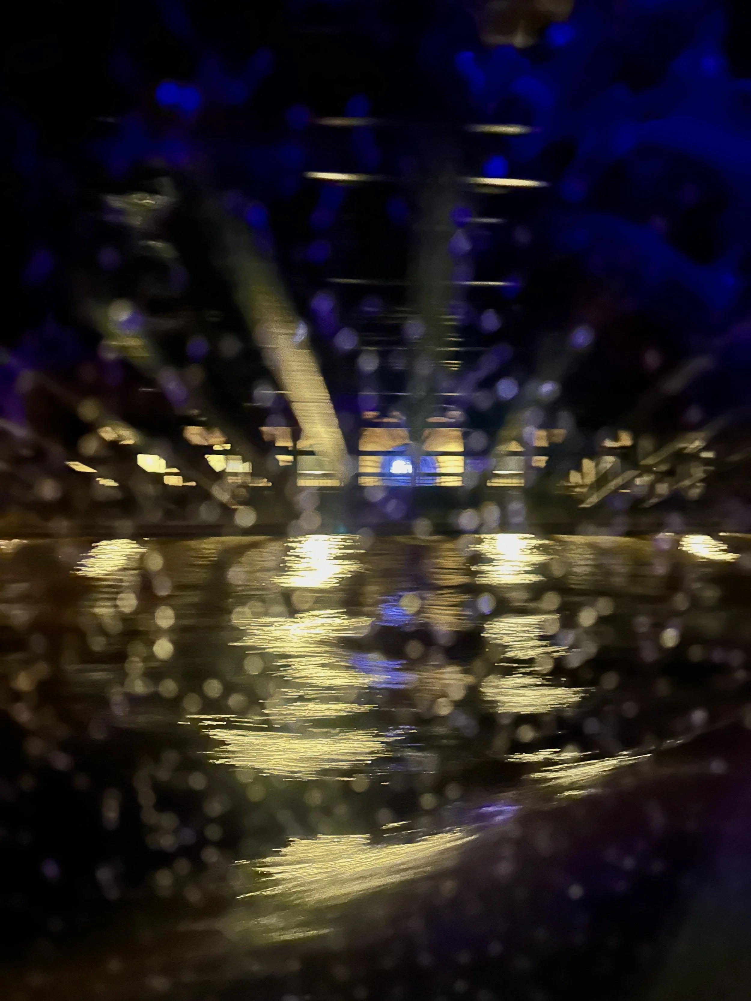 Nighttime photo of a bridge over water, taken through raindrops on a window with reflections of lights on the water.