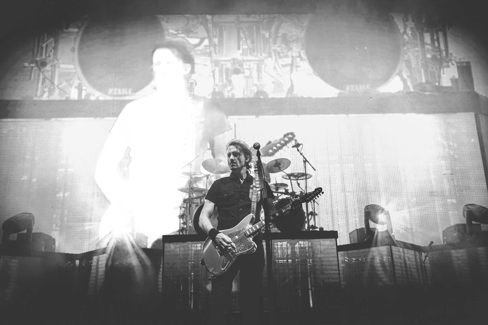 Black and white photo of a musician performing on stage, playing an electric guitar with a drum set in the background and large screens displaying visuals behind him.