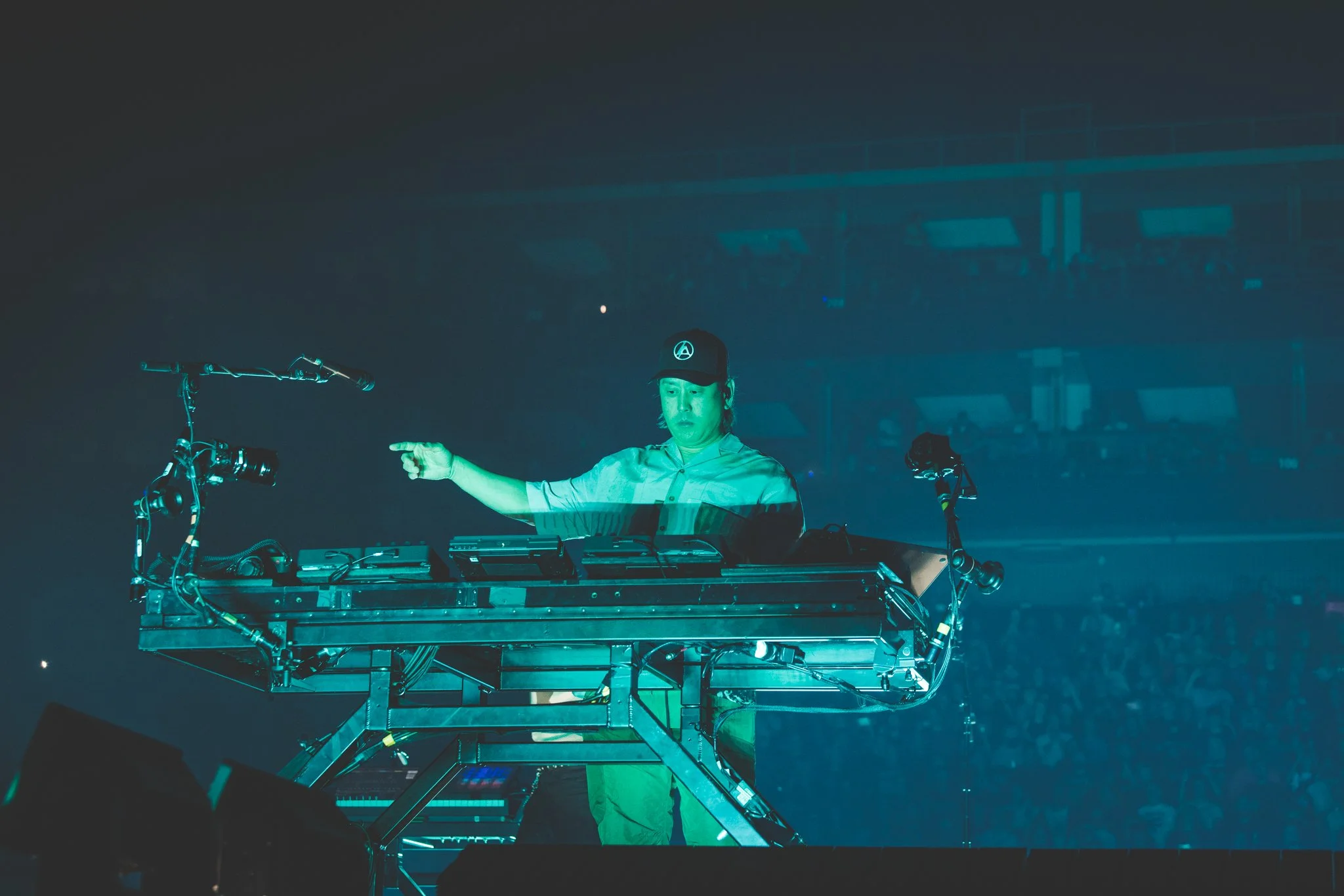 DJ performing on stage at a concert, pointing with his right hand. He wears a hat and sports a serious expression, surrounded by DJ equipment in a dark, crowded venue.