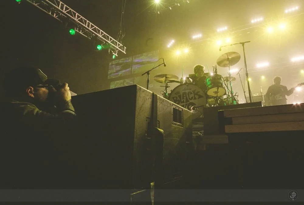 A concert scene with a drummer behind a drum set that has the band's name, "The Black" visible on the bass drum, on a brightly lit stage with yellow and green lights, and camera crew or photographers in the foreground.