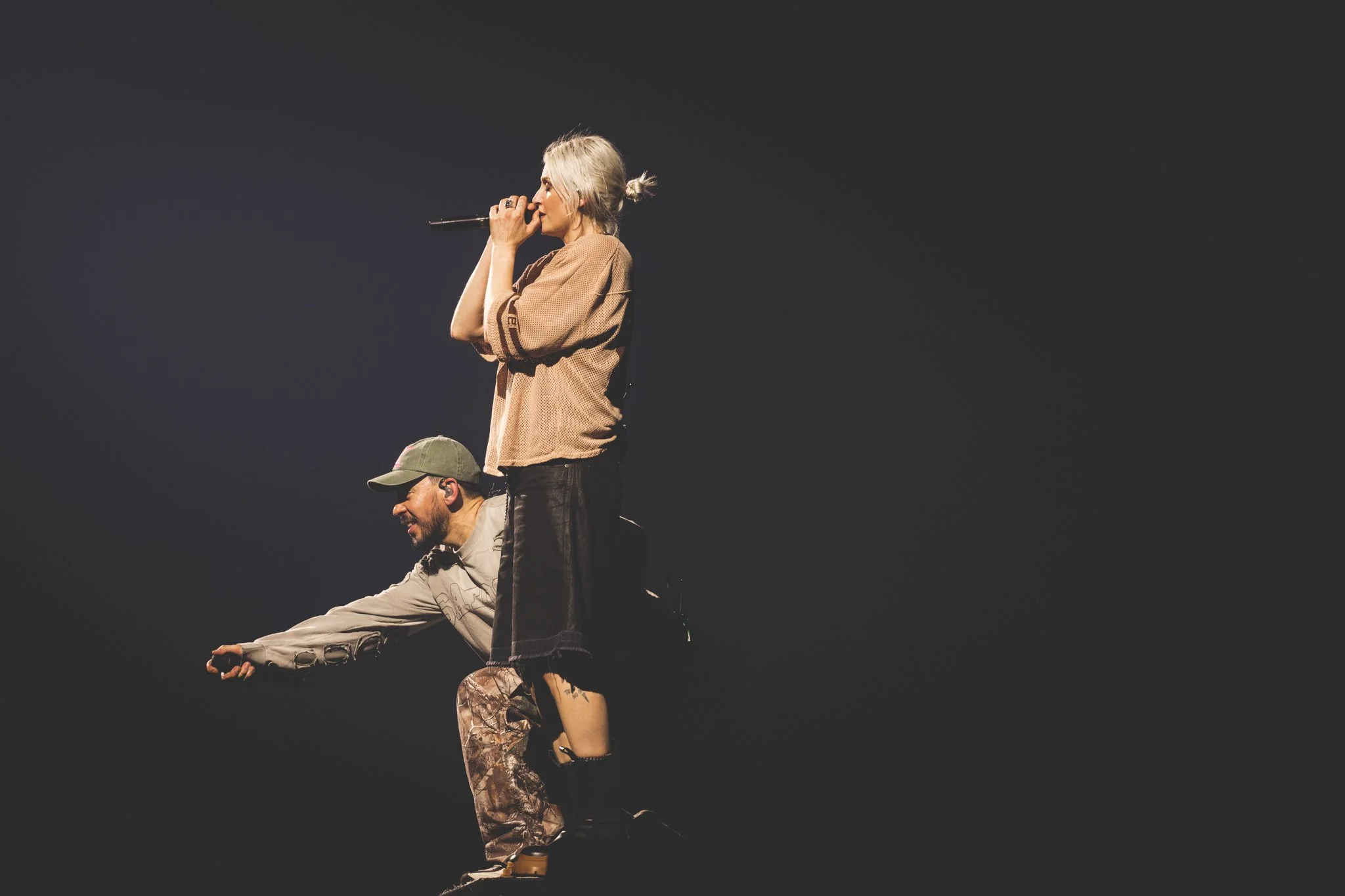 A man and a woman performing on stage, with the woman singing into a microphone and the man in camouflage pants, wearing a cap, supporting her by holding her leg. The background is dark and the stage is dimly lit.