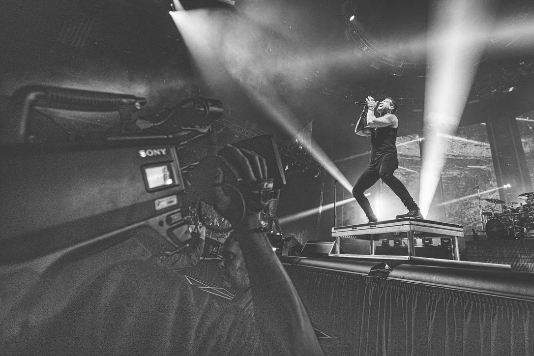 A musician performs on stage at a concert, standing on a platform with lights shining behind him, while a camera operator records him in the foreground.
