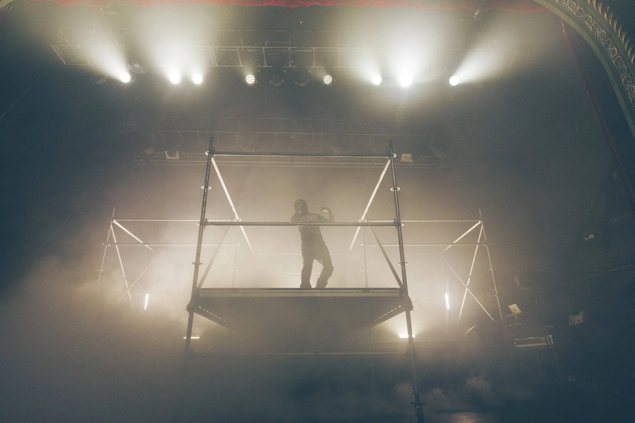 Person standing on a high scaffold platform with bright stage lights above, surrounded by fog or smoke, inside a theater or concert hall.