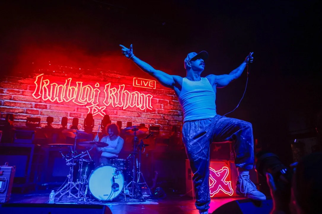 Performer with outstretched arms on stage at a rock concert, neon sign reads "Karen O Live" in the background, drummer playing behind him, stage lights illuminate the scene.