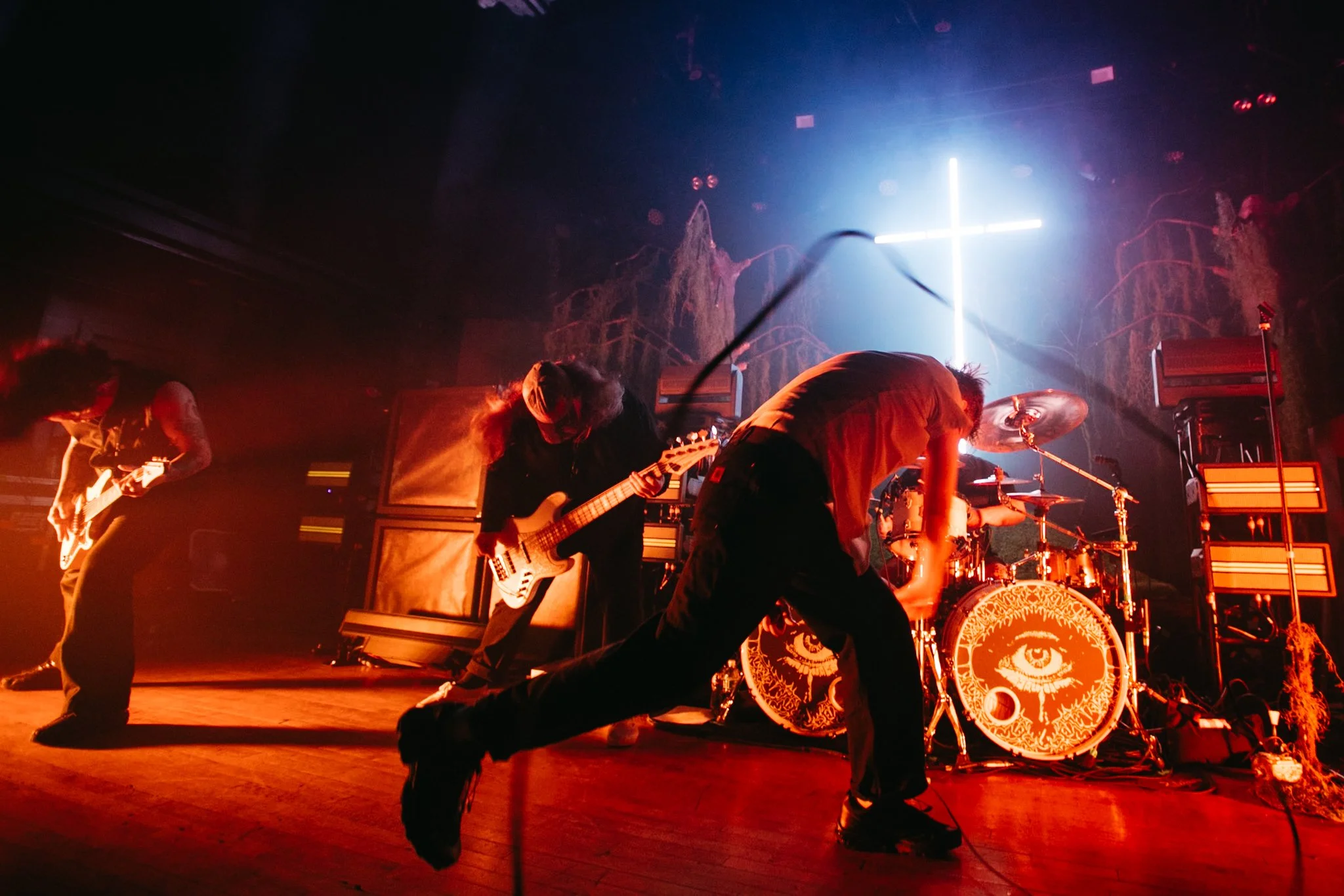 A band performing live on stage with a neon cross in the background and an eye symbol on the bass drum, with dynamic lighting and smoke effects.