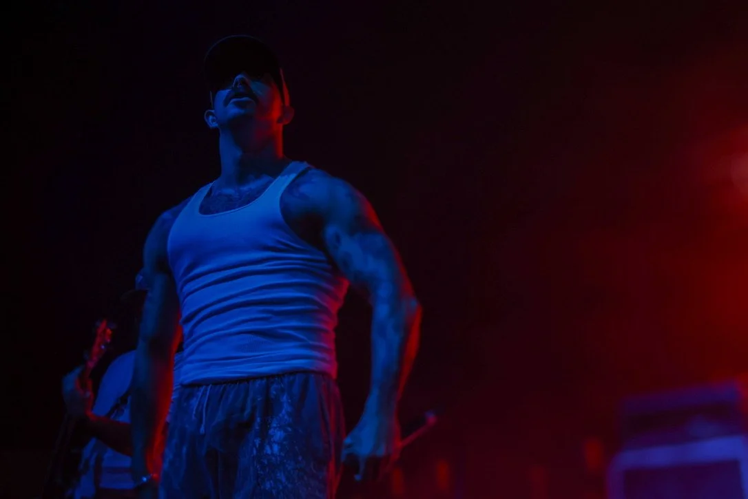 A man wearing a white tank top and shorts, with tattoos on his arm, stands in dark lighting with red and blue lights, wearing a cap and sunglasses.