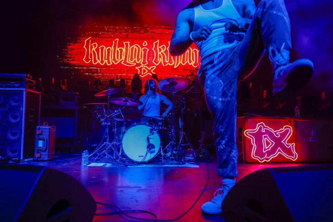 Musicians performing on stage with a drummer, a person in muscle shirt, and a person kicking with neon signs and red and blue lighting.