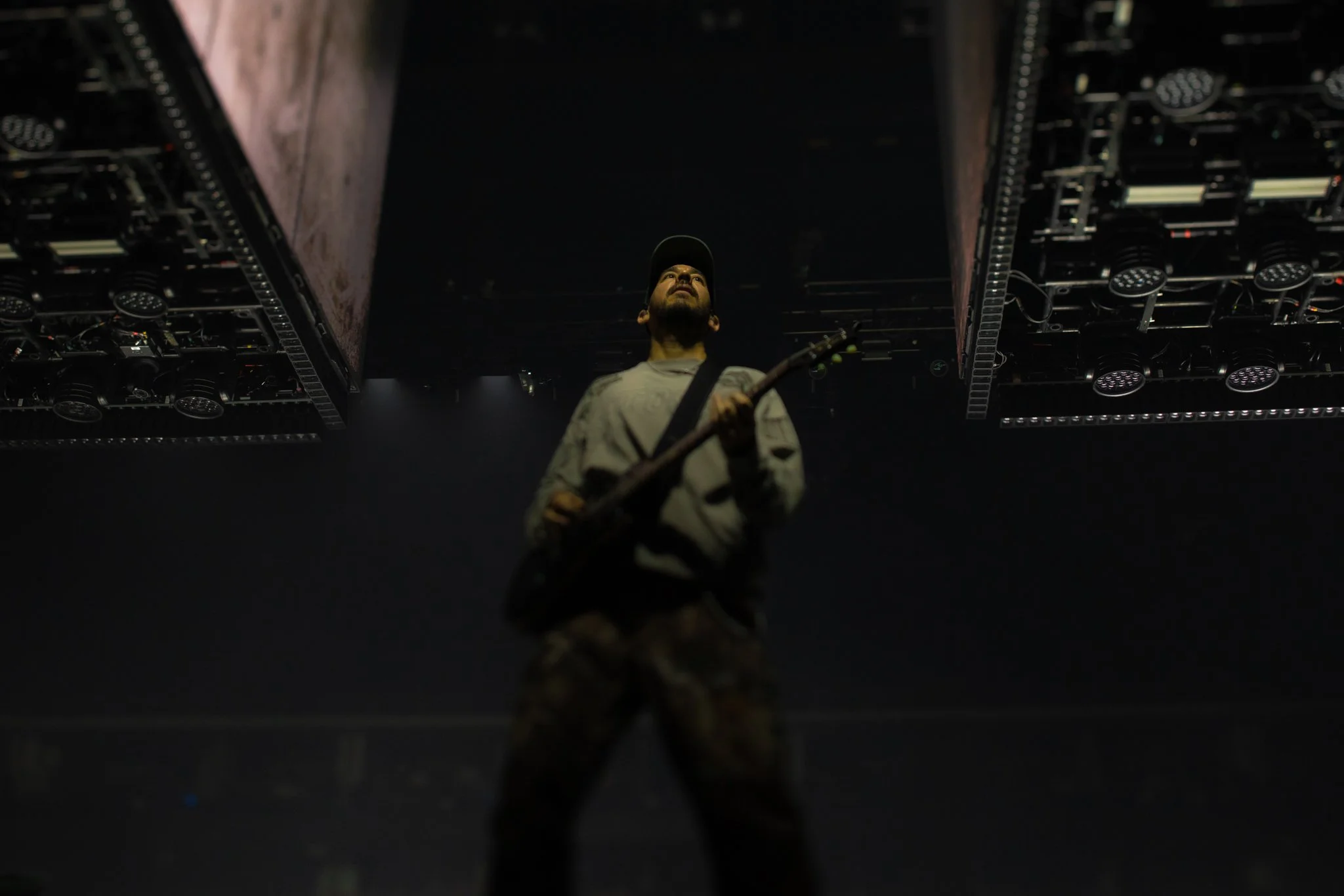 A man holding a guitar on stage under dark lighting, with stage equipment overhead.