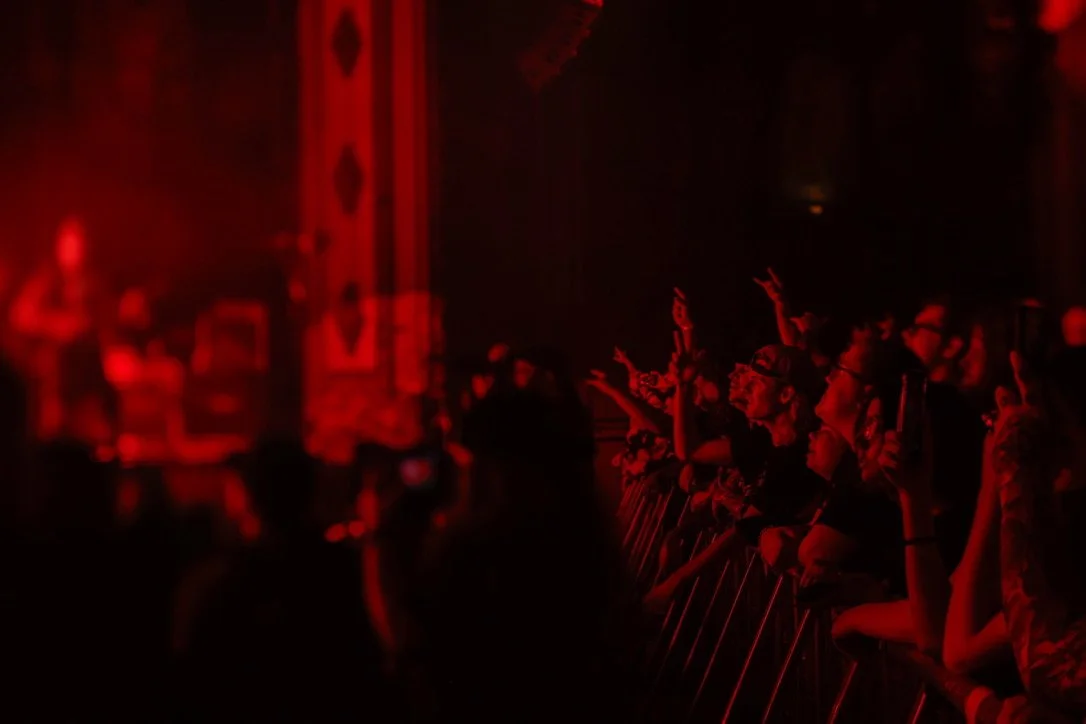 Audience at concert or event illuminated by red lighting, some individuals raising their hands, facing the stage.