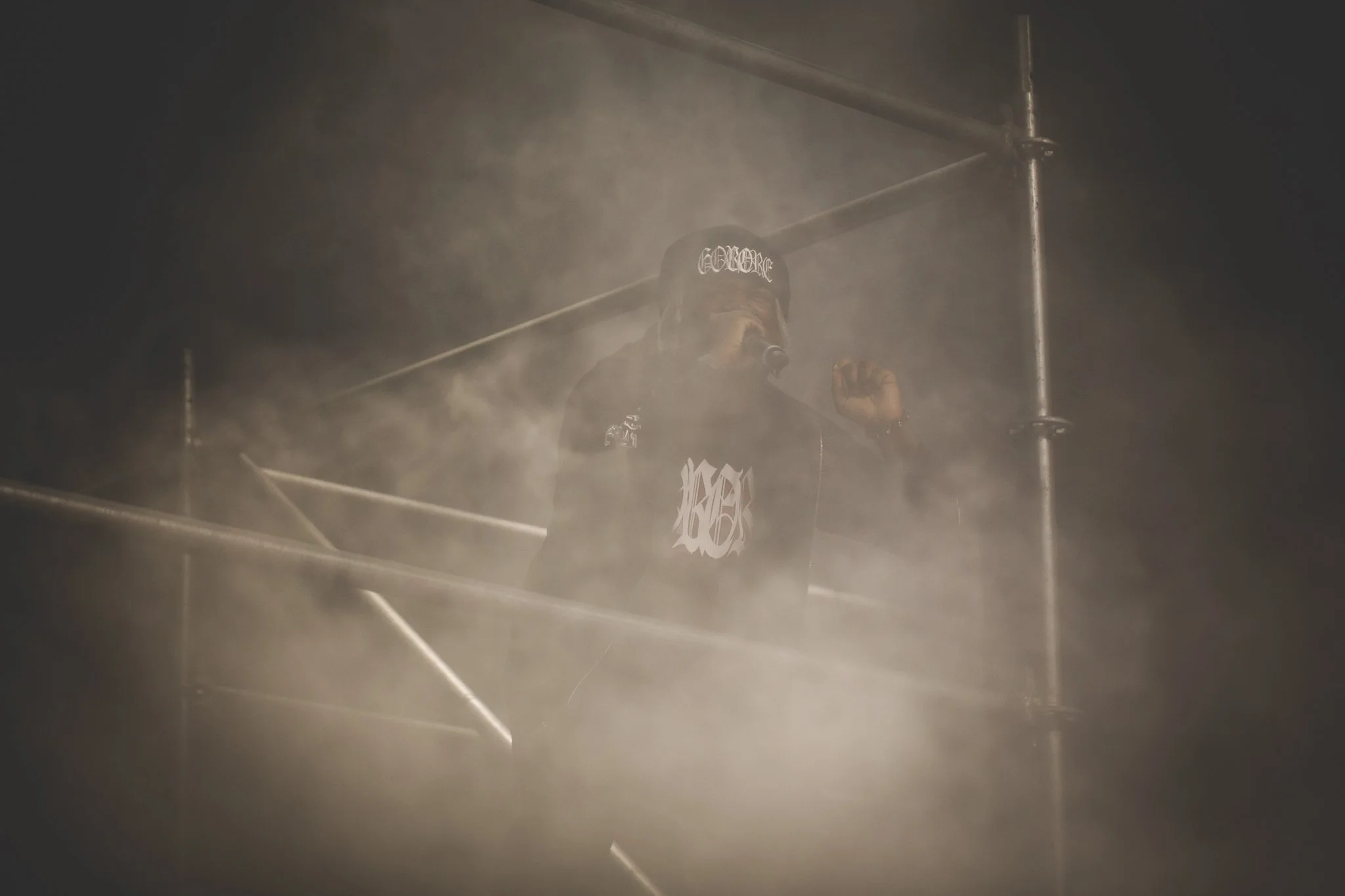 A performer on stage surrounded by fog or smoke, wearing a black beanie and hoodie, holding a microphone, with metal scaffolding in the background.