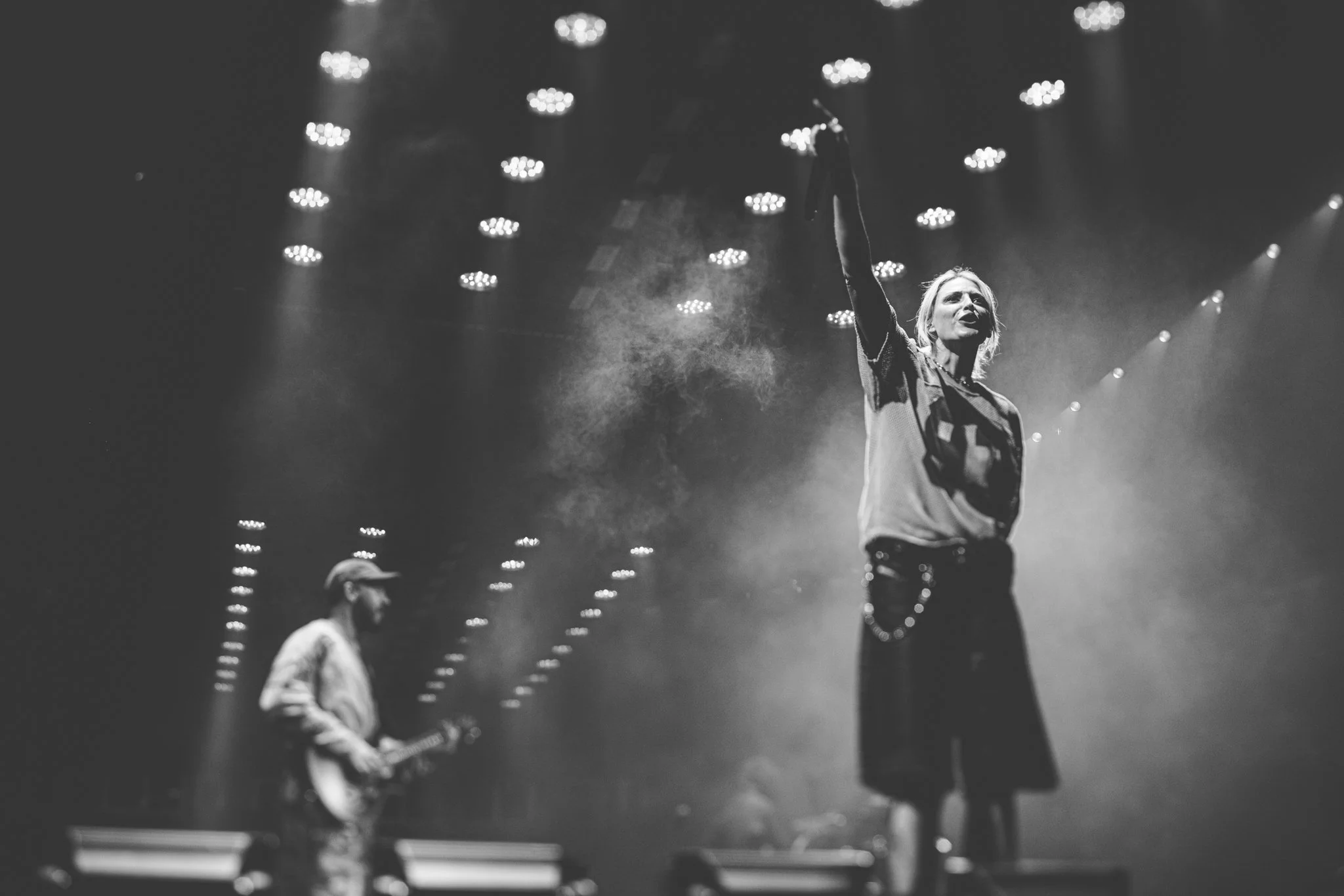 A vocalist on stage holding a microphone and raising her hand, with a guitarist playing in the background, under stage lights in a black and white photo.