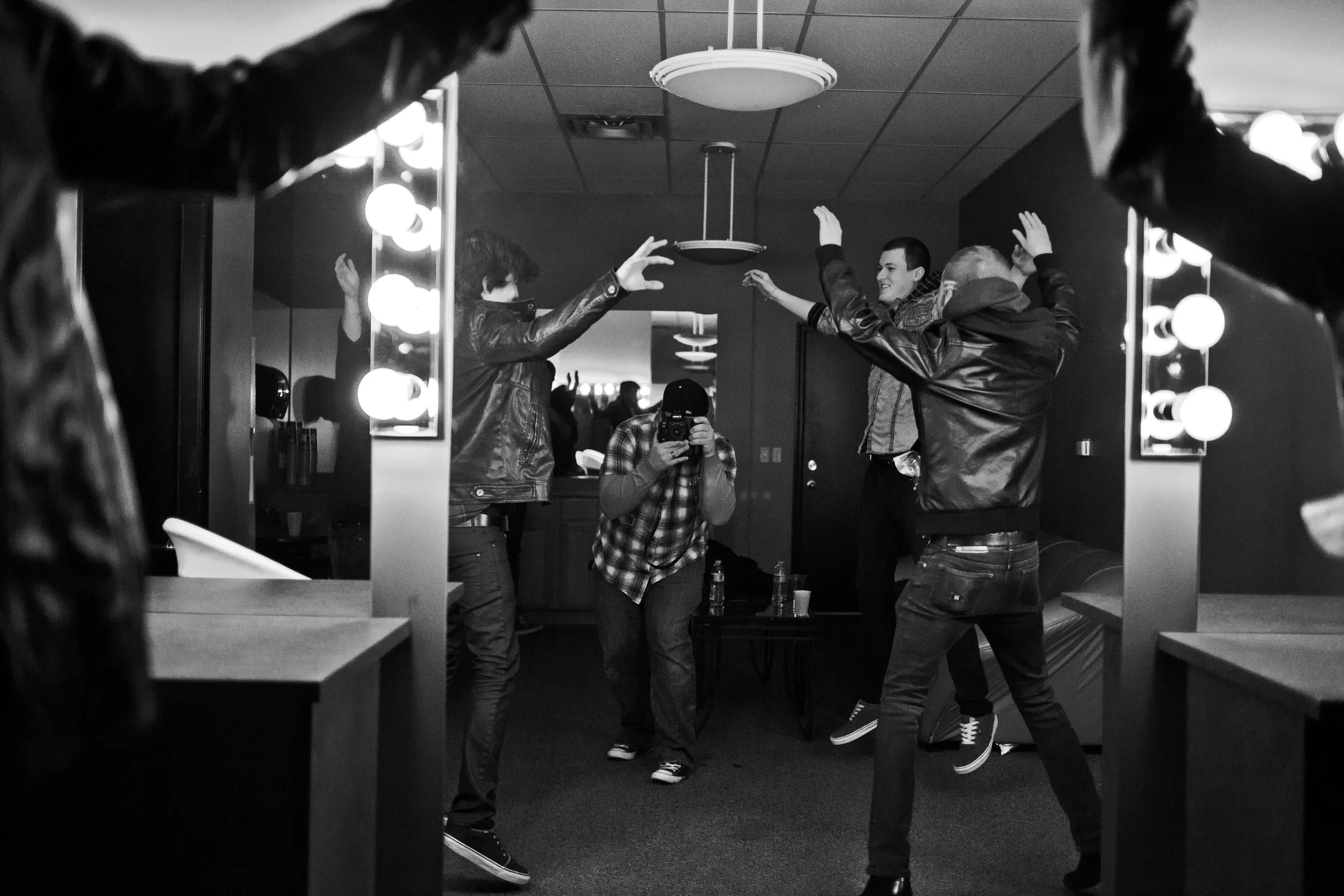 People dancing and enjoying themselves in a backstage dressing room, reflected in a large mirror with bright vanity lights, in black and white.