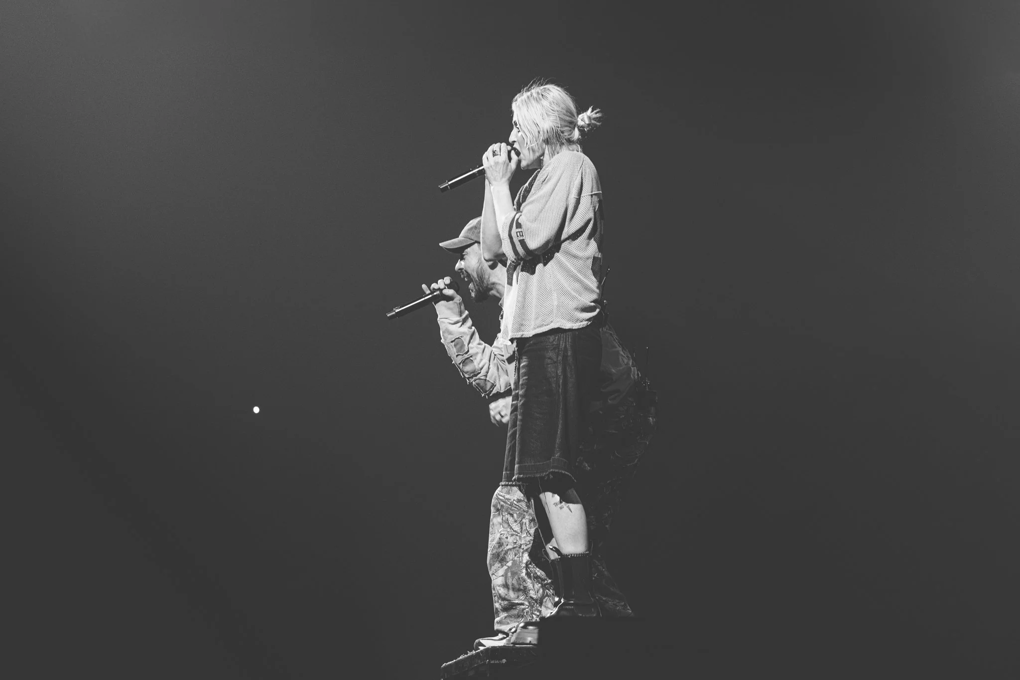 Black and white photo of a man and woman singing into microphones on stage, with the woman standing and the man kneeling. The woman has blonde hair tied back, wearing a loose top and shorts, while the man has a beard, wearing a cap, jacket, and patterned pants. The background is dark with a small bright spot visible.