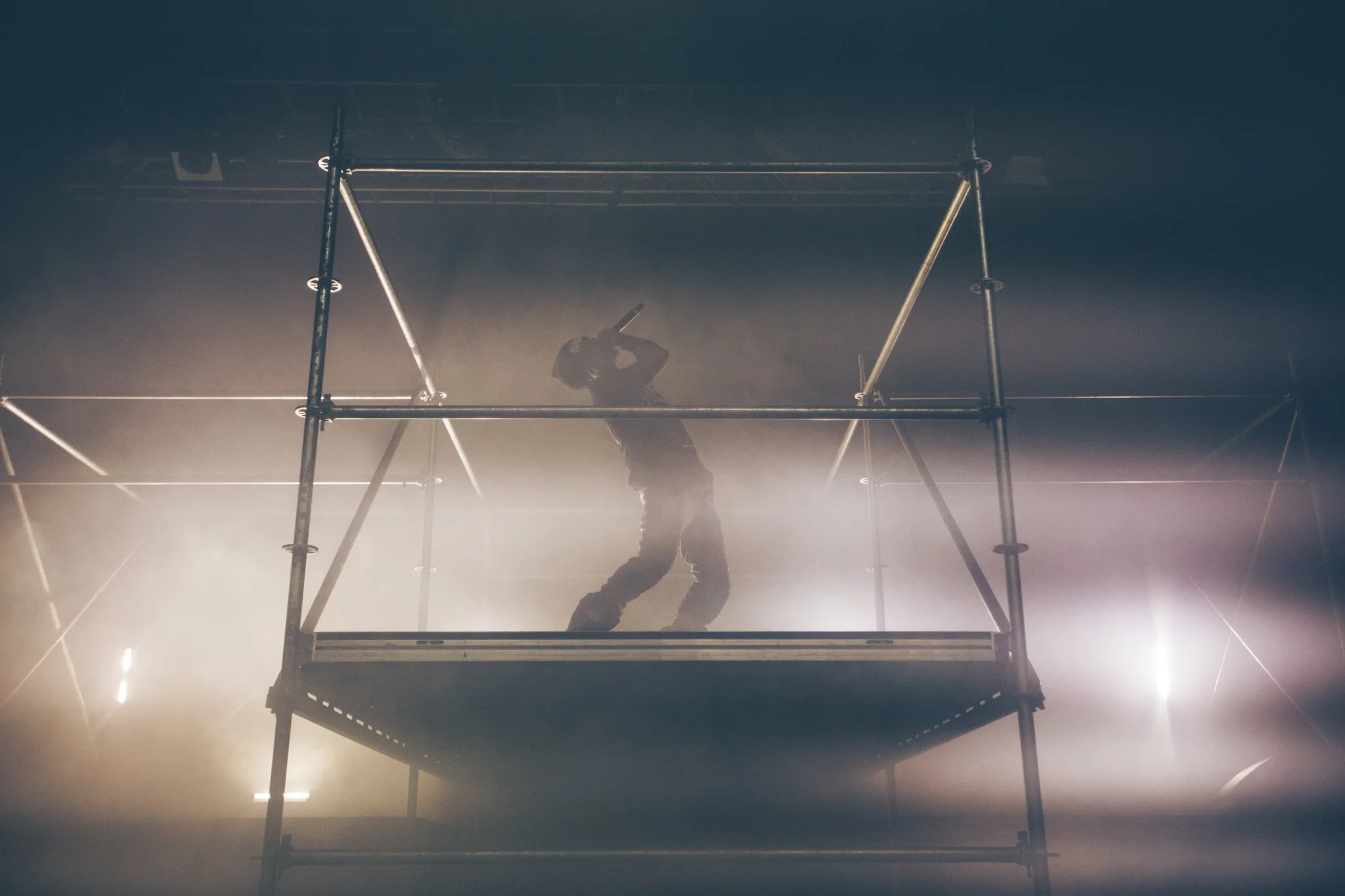 A performer standing on a metal platform surrounded by fog, holding a microphone near their mouth.