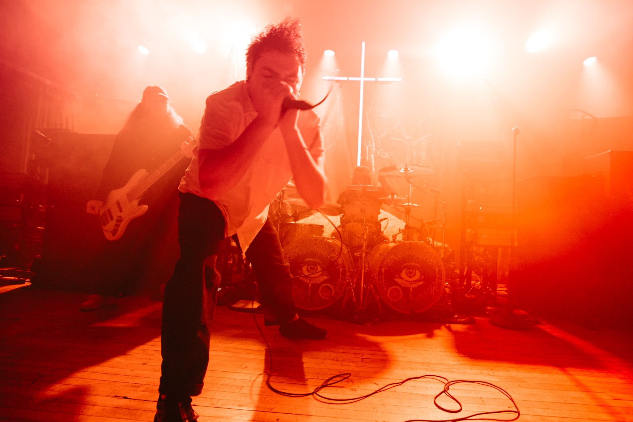 A band performing on stage with red lighting, featuring a singer holding a microphone, a bass guitarist, and a drummer with eye designs on the drums, and a neon cross in the background.