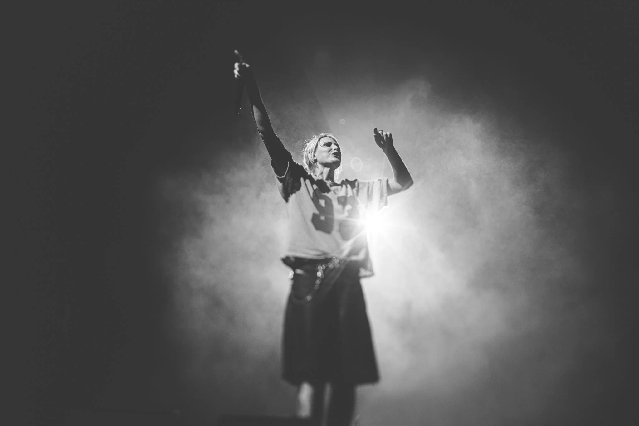 Black and white photo of a woman on stage with arms raised, illuminated from behind, creating a dramatic lighting effect.