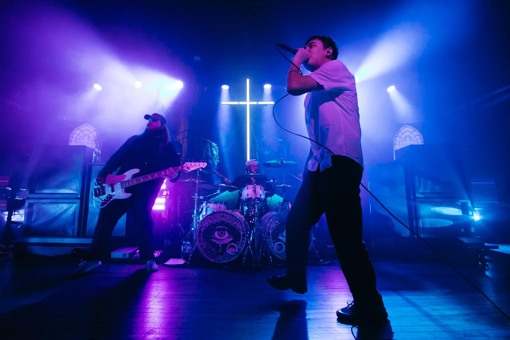 Band performing on stage with purple and blue lighting, a large illuminated cross in the background, and a drum kit with eye and moon symbols on the bass drums.