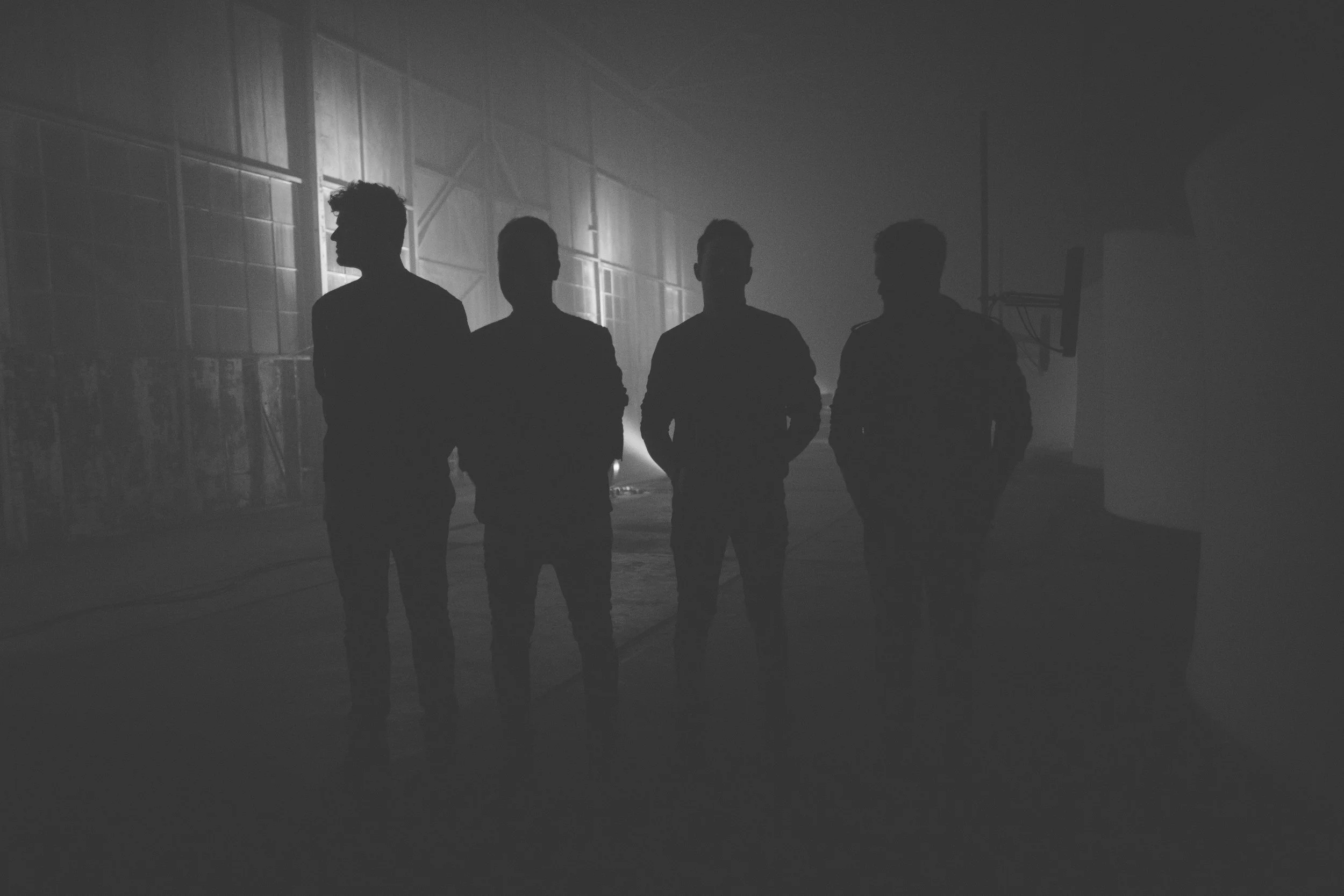 Four people stand in a dim, foggy environment with a warehouse-like background, silhouetted against a light source behind them.