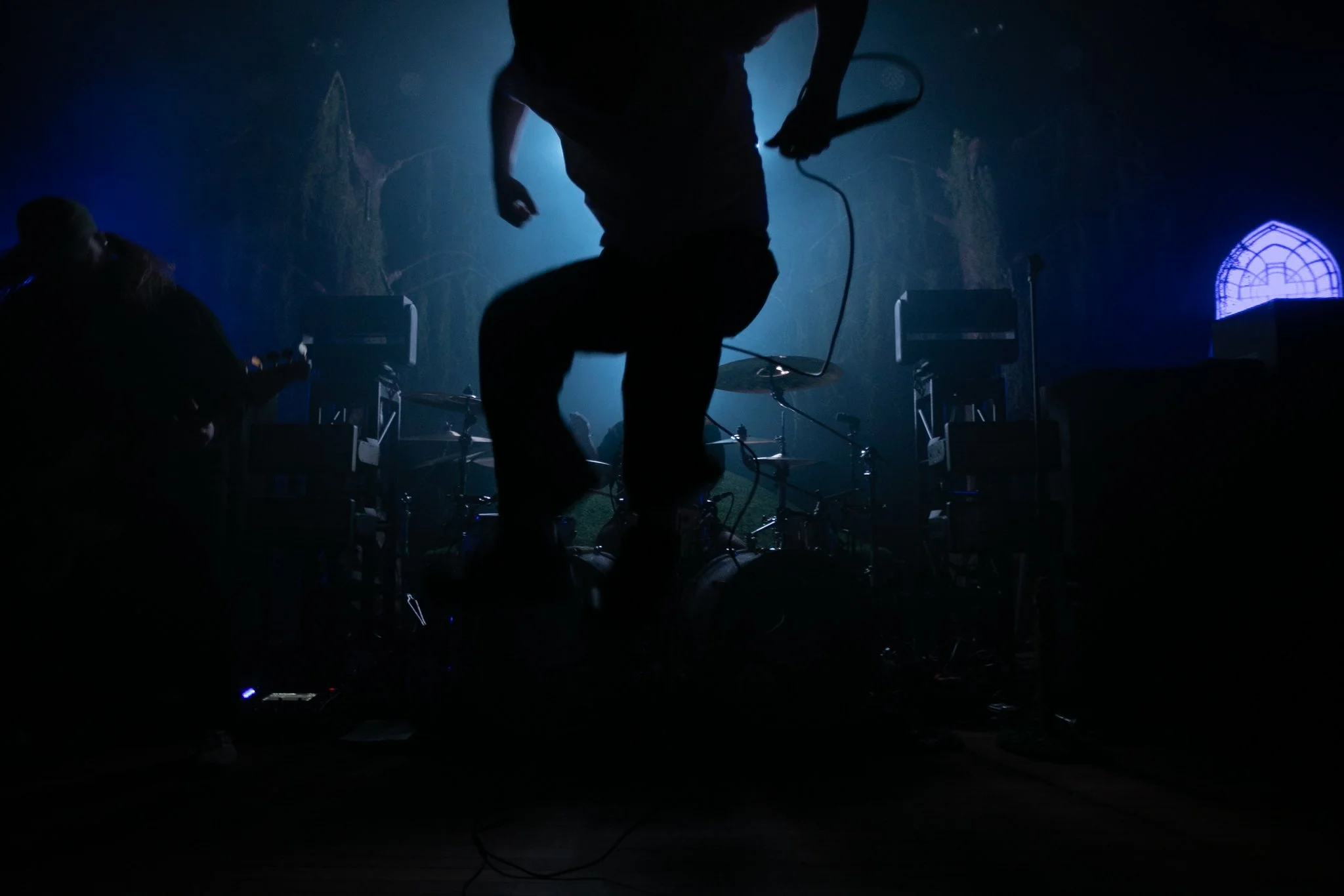 A musician jumping on stage during a concert with musical instruments and chairs in the background, illuminated by blue lighting.