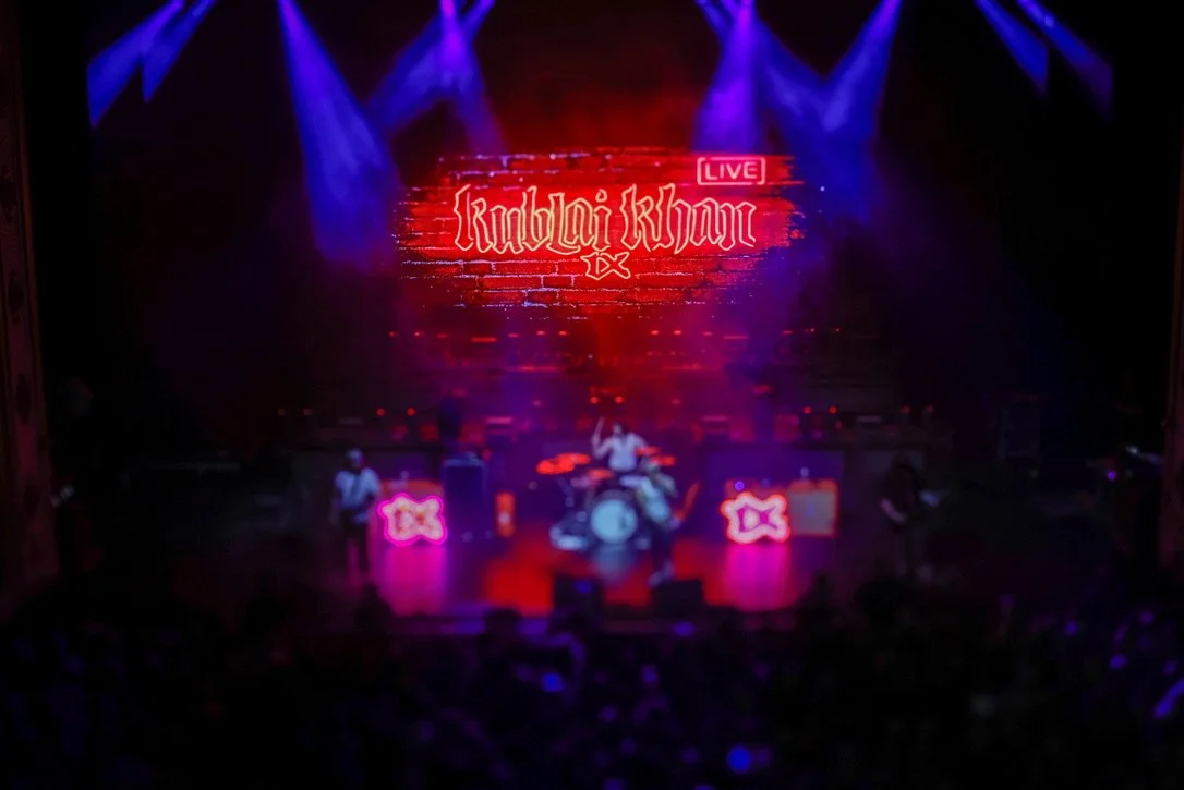 Live music performance on stage with band, large digital screen with red neon text 'Khalil Kherr', purple stage lighting, and audience in front.