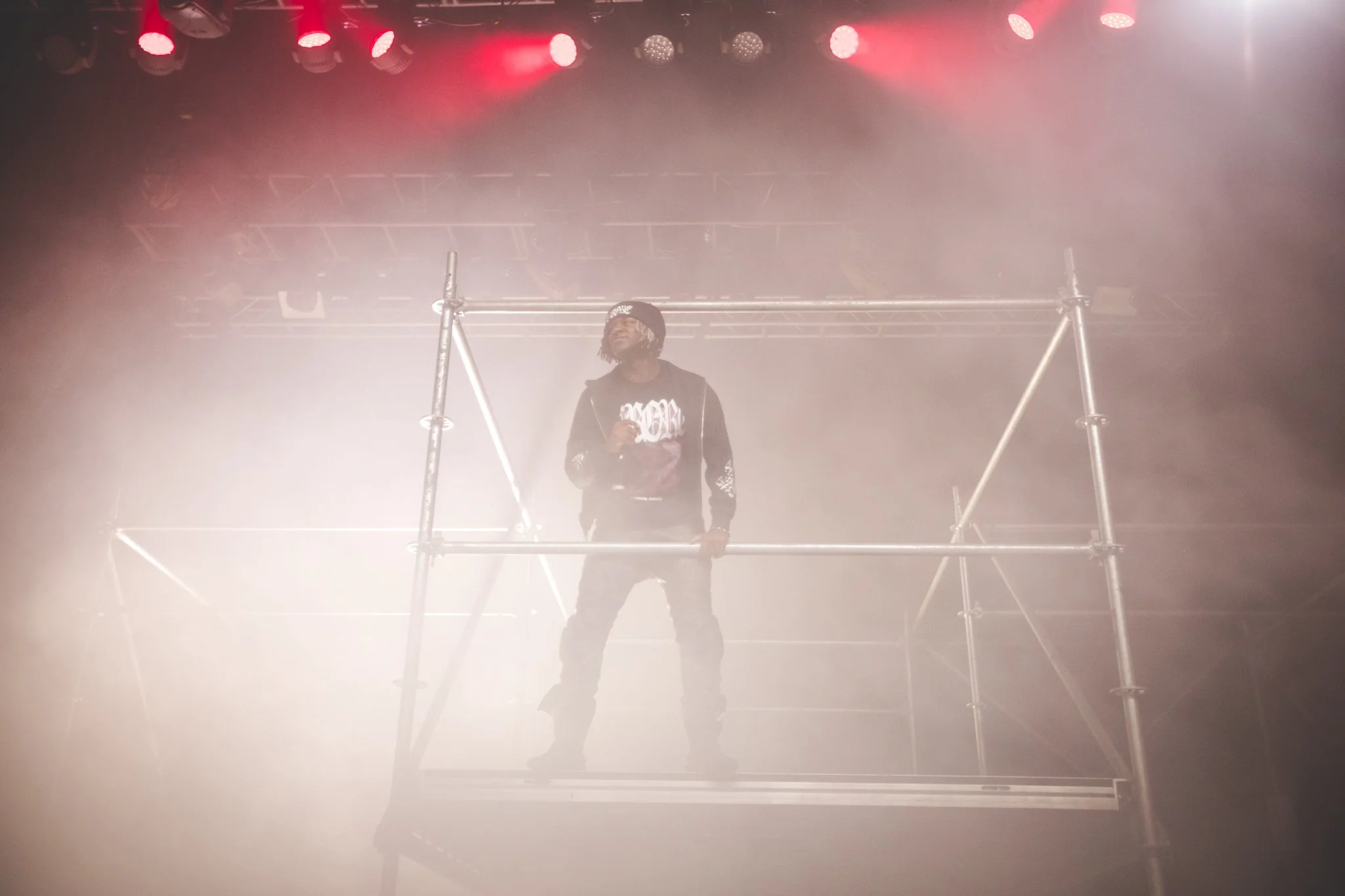A performer standing on a metal scaffold stage surrounded by fog and red stage lights.